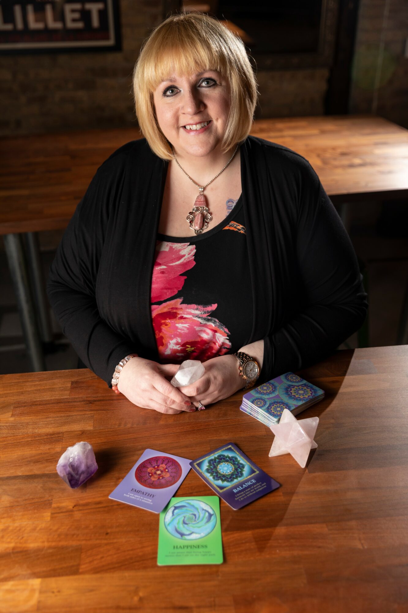 Woman with blonde hair smiling, sitting at a table with tarot cards and crystals, in a dimly lit room.