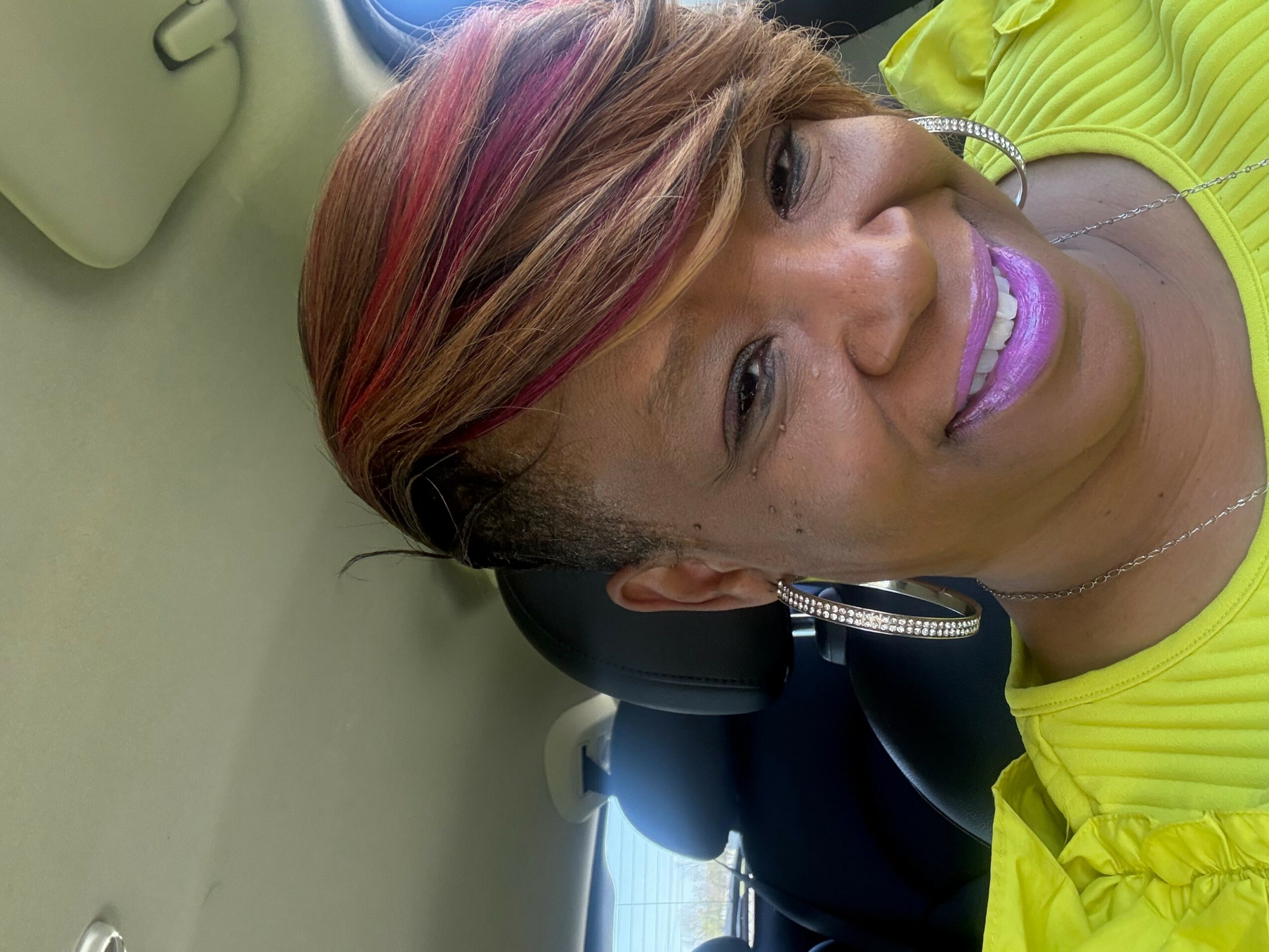 Woman with colorful hair smiling, wearing a yellow top, sitting in a car, with a light green purse nearby.