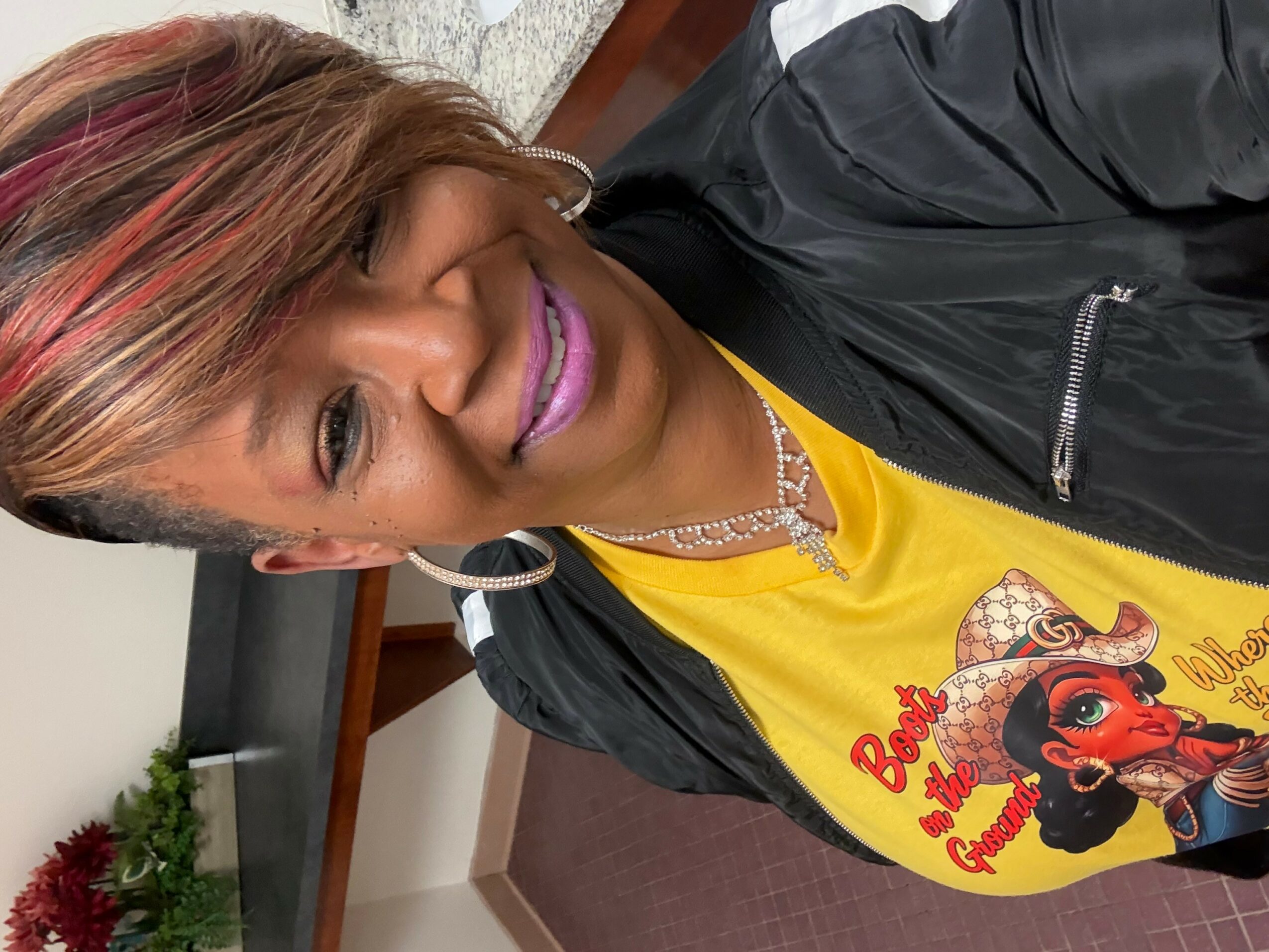 Woman with short hair, wearing a yellow shirt with a cartoon character, black jacket, and jewelry, smiling indoors.