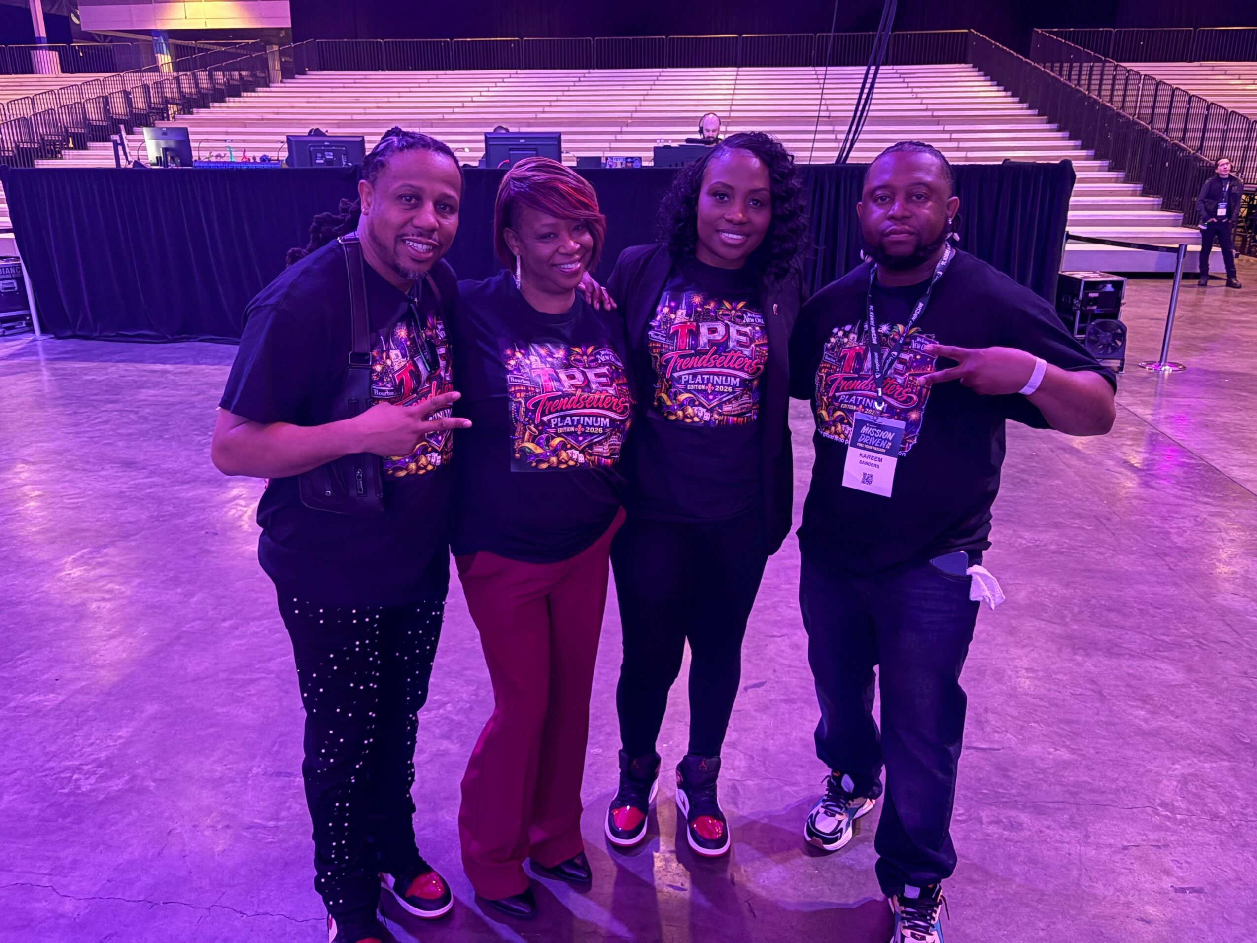 Four people standing together in an indoor venue, smiling, with a stage and seating in the background.