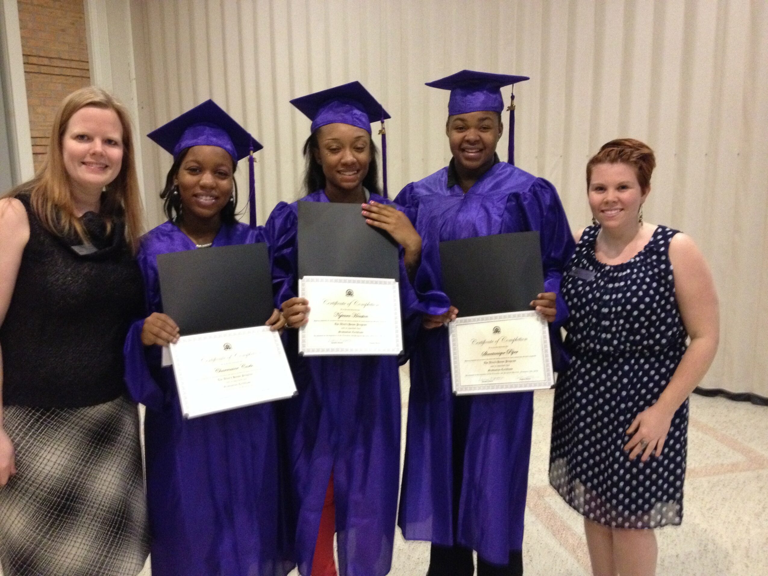 Four graduates in purple caps and gowns holding diplomas, two women in casual dresses stand beside them, smiling.
