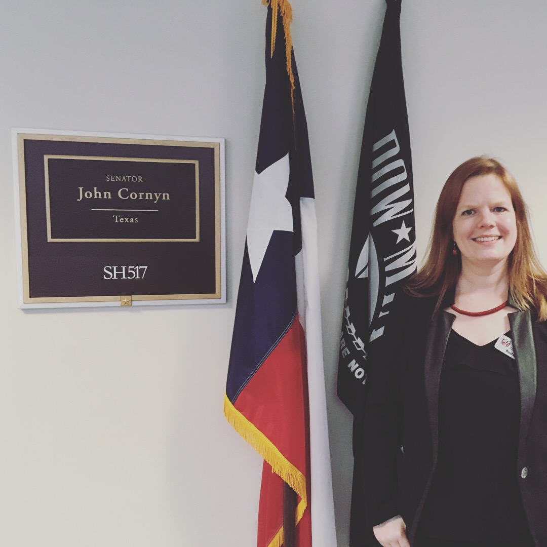 Woman with shoulder-length hair smiling, standing next to flags and a sign for Senator John Cornyn, Texas.