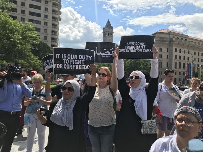 Group of people protesting outdoors, holding signs, with a cityscape and clock tower in the background.