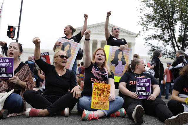Group of women protesting, holding signs and raising fists, sitting on the ground outdoors, with a building and trees in background.