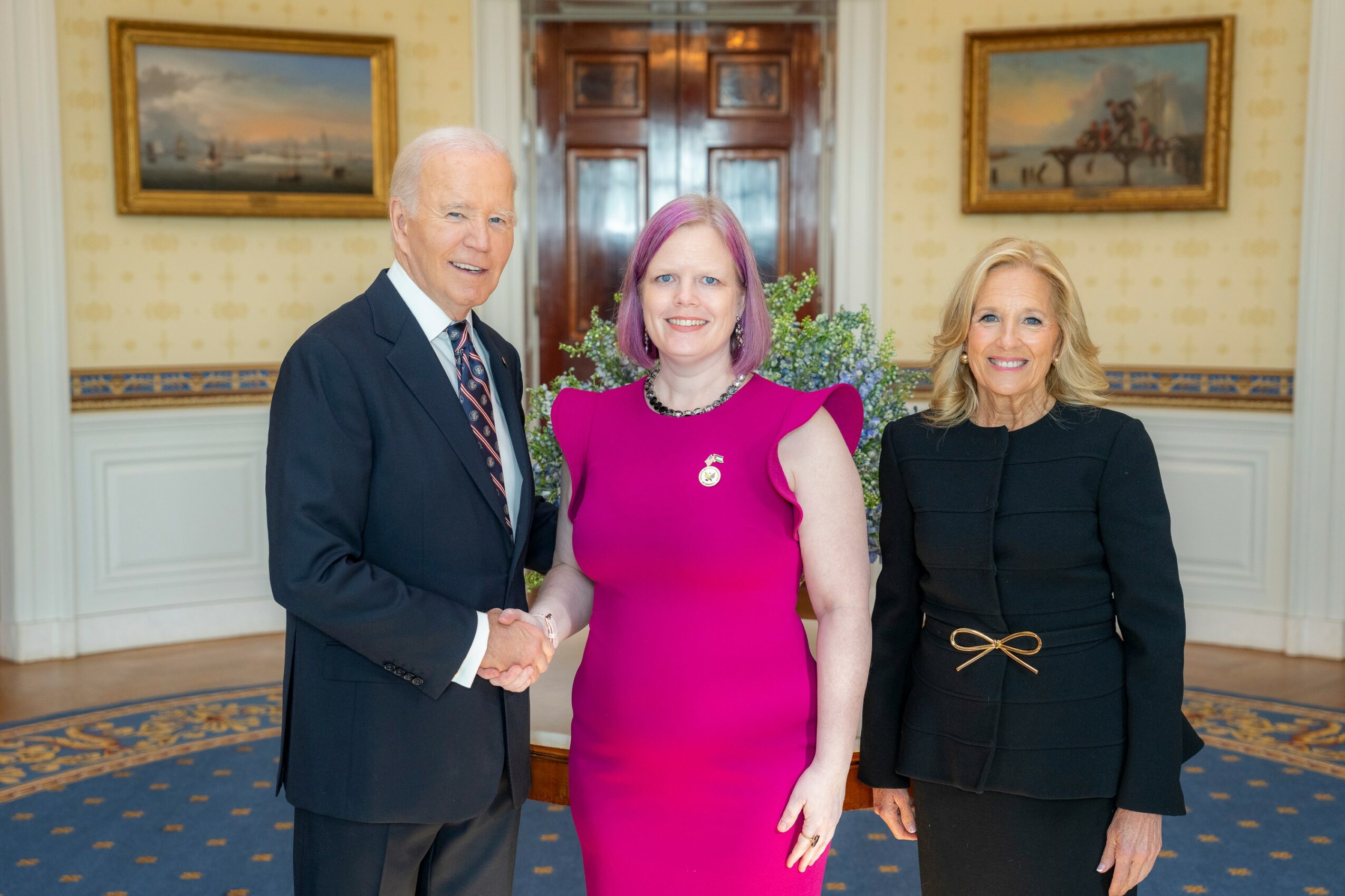 Three people standing in a room with paintings, smiling, with a man shaking hands with a woman in a pink dress, and another woman nearby.
