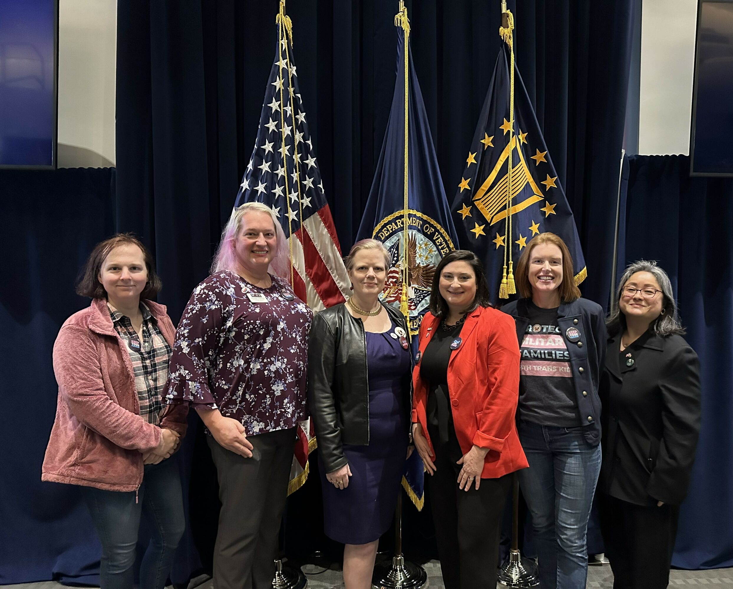 Six women stand in front of American and state flags on a stage, smiling for a group photo.