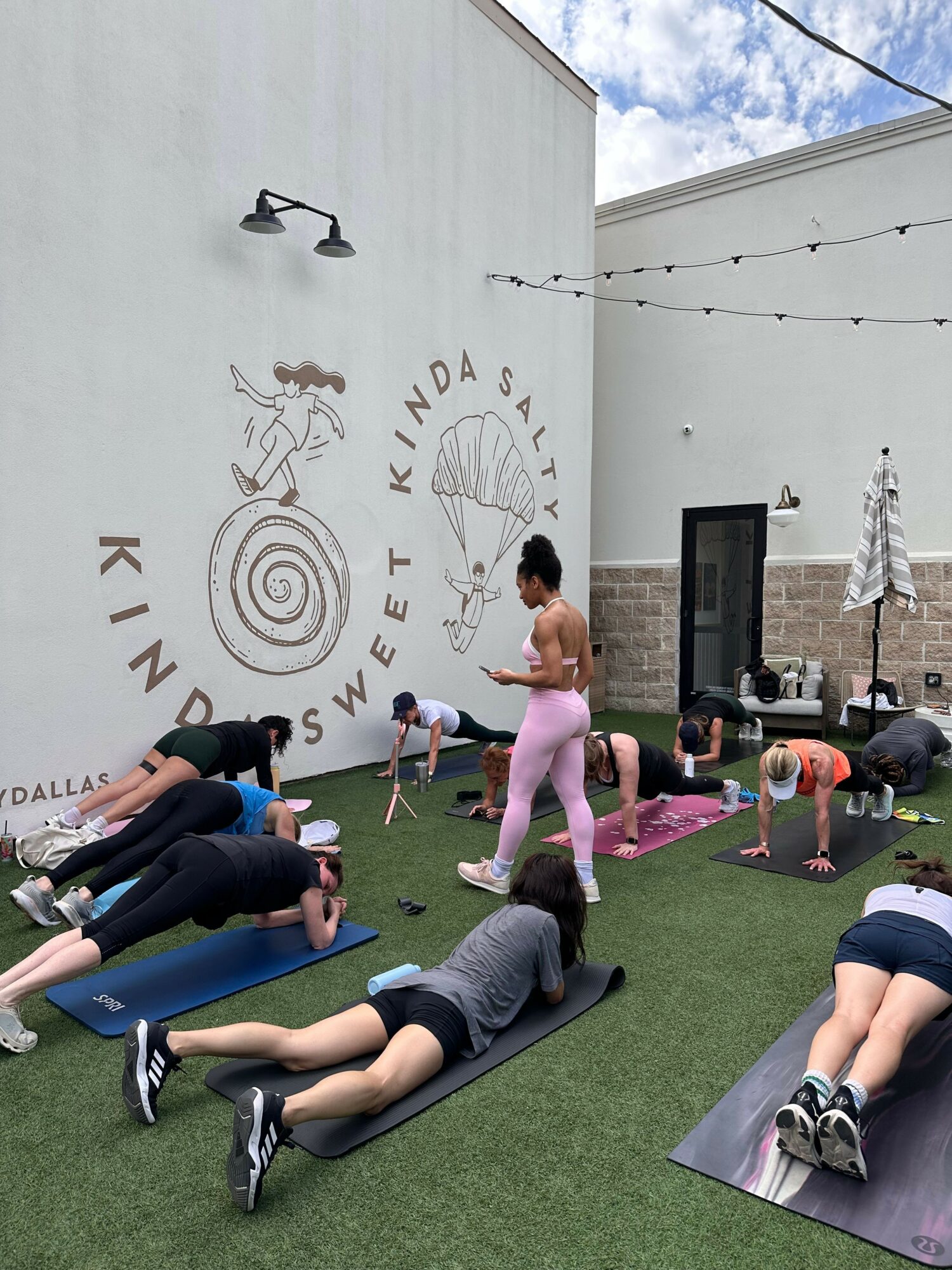 Children and adults gather outdoors near a white wall with a logo and text, some sitting on mats, others standing or walking.