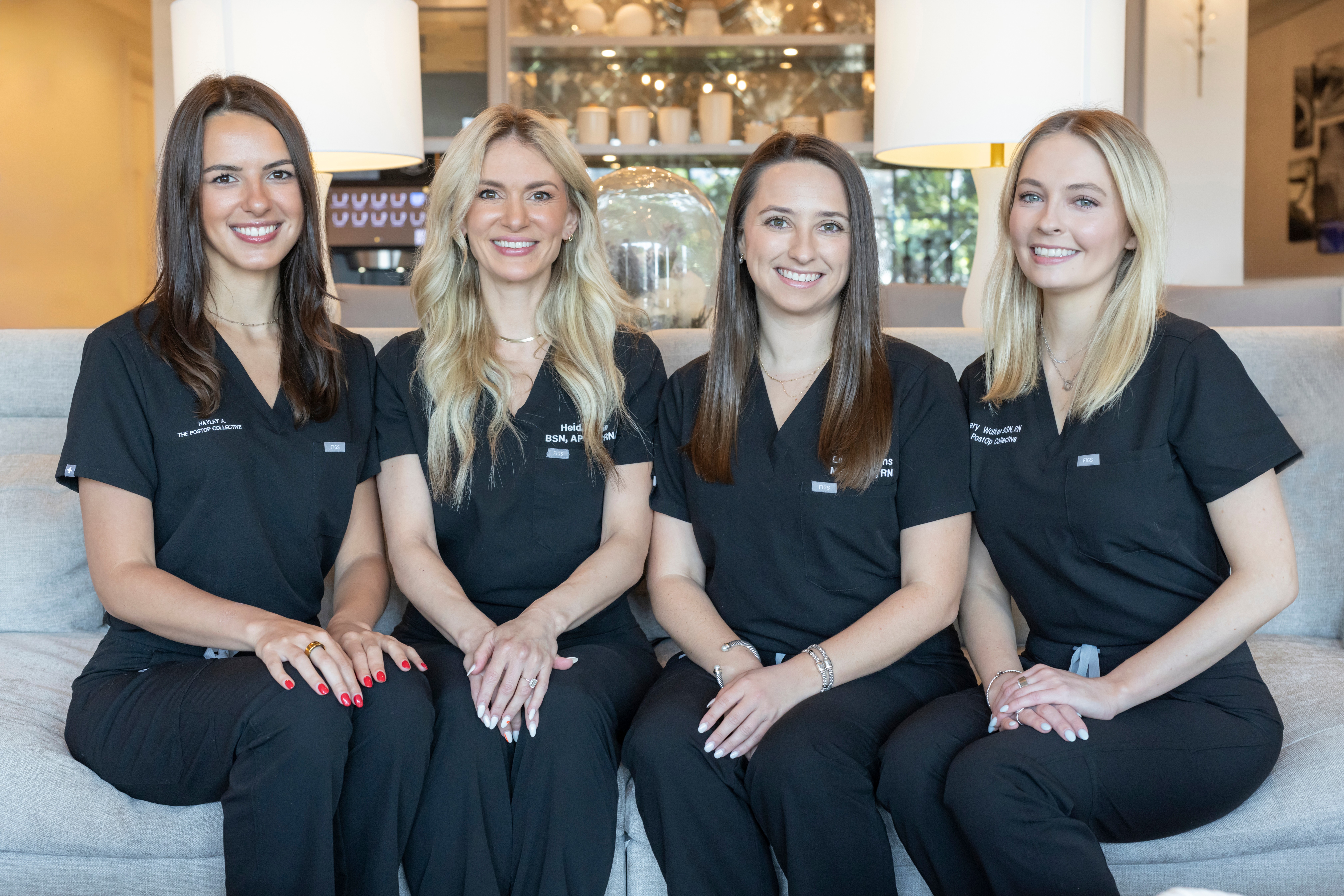Four women in black uniforms sitting on a sofa, smiling, in a well-lit room with shelves and decor behind them.