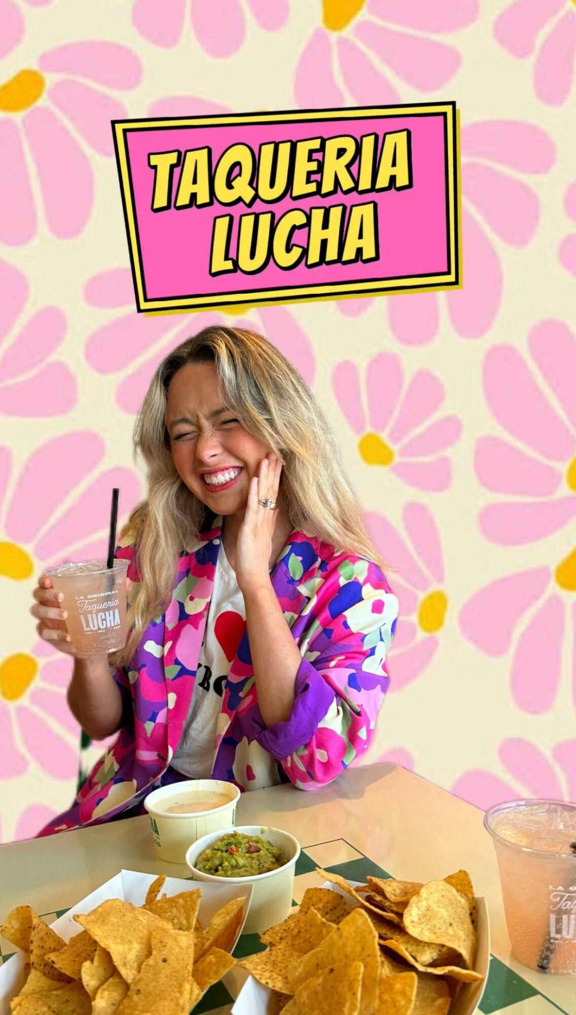 Woman smiling with a drink at a taco stand, colorful background, food on table, cheerful atmosphere.