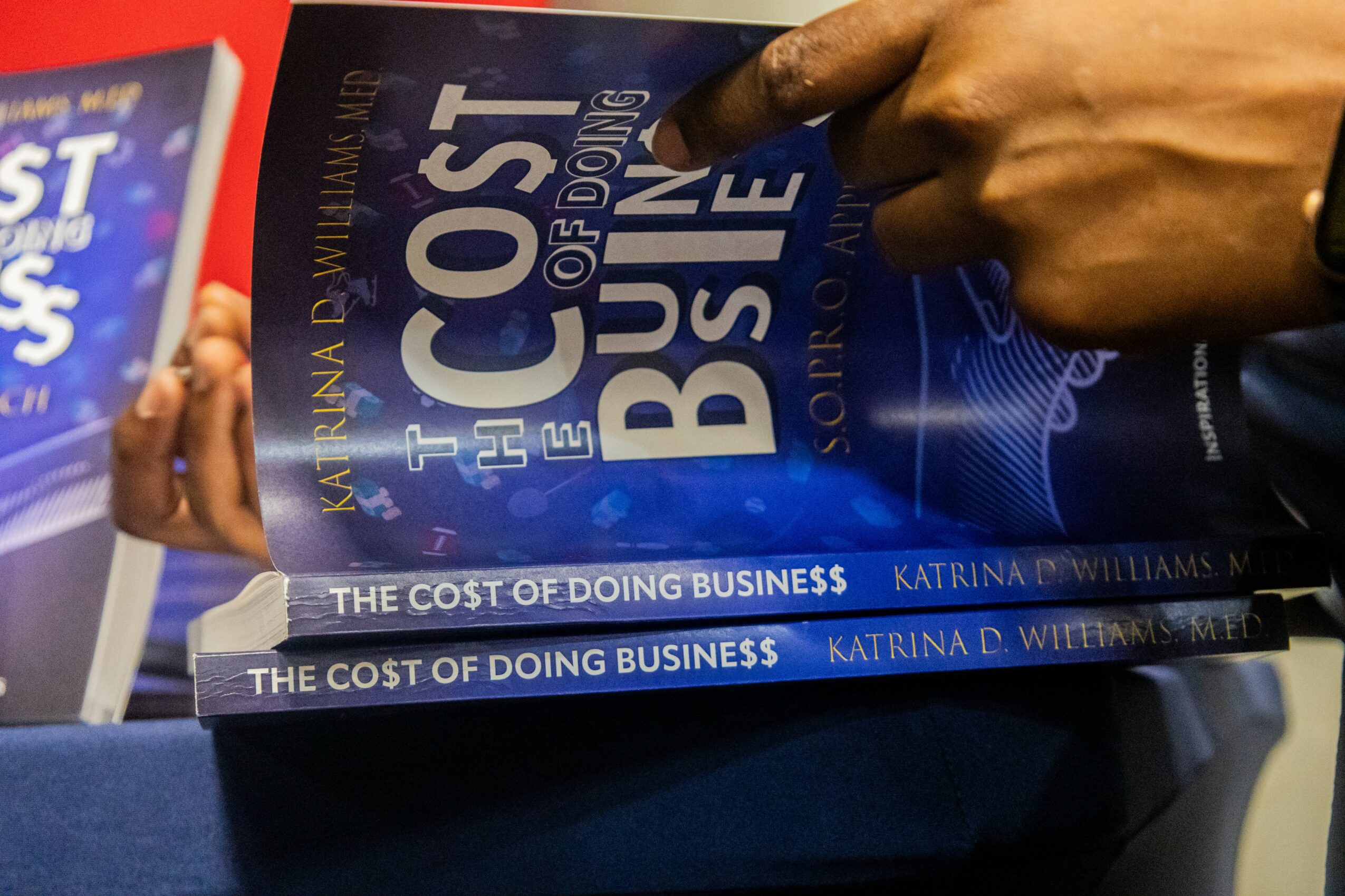 Person holding two copies of a book titled 'The Cost of Doing Business' by Katrina Williams, with a blue cover and white text.