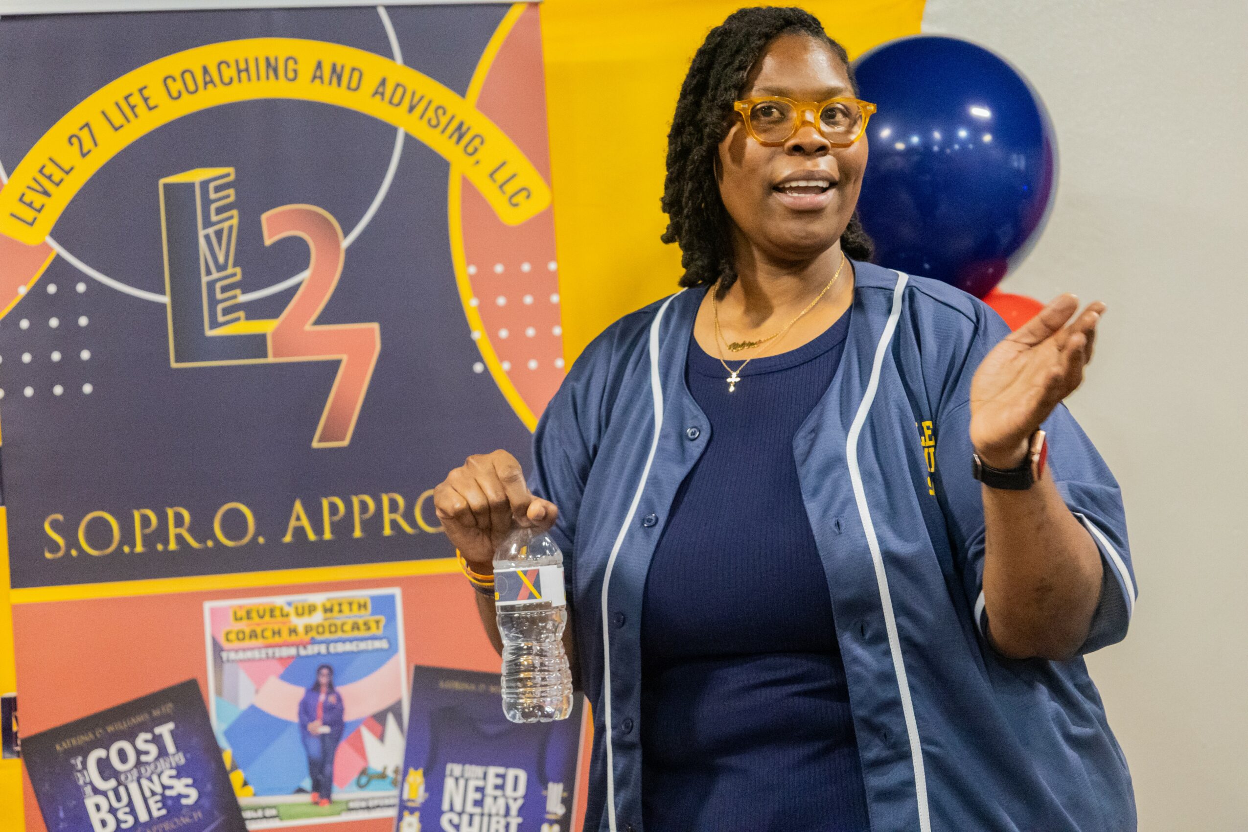 Woman speaking in front of a banner with coaching and approval text, holding a water bottle, with blue and yellow background elements.