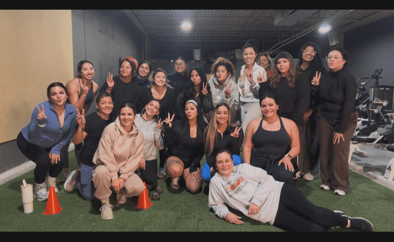 Group of women and one man posing indoors, some making peace signs, with exercise equipment in background.