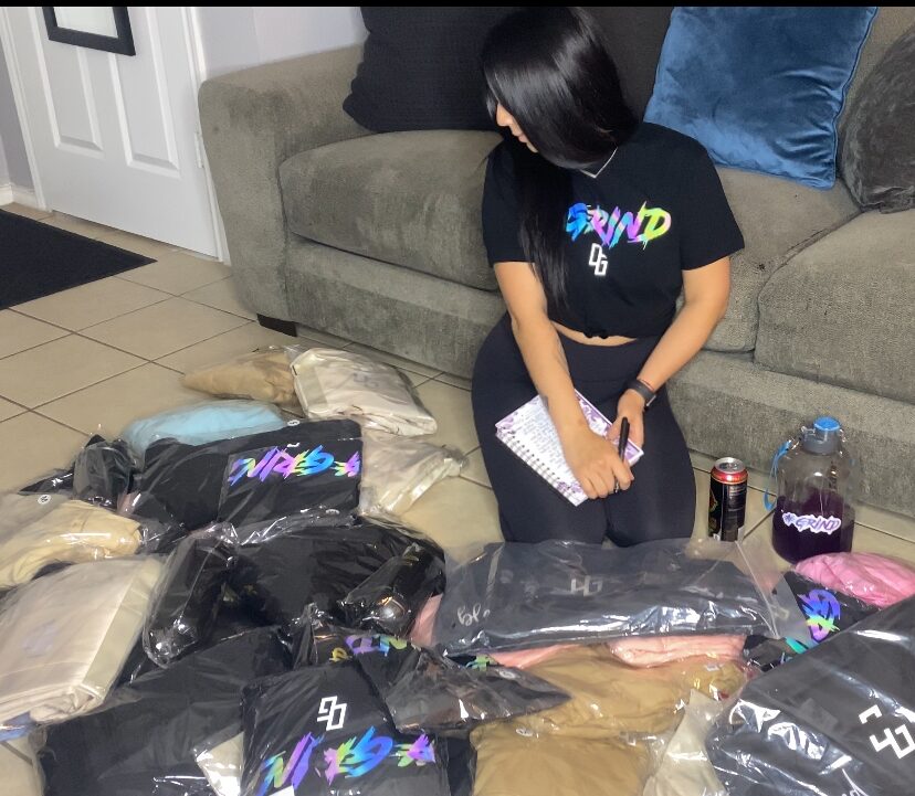 Person with black hair wearing a black T-shirt with colorful text, sitting on a gray couch, surrounded by plastic bags and items on the floor.