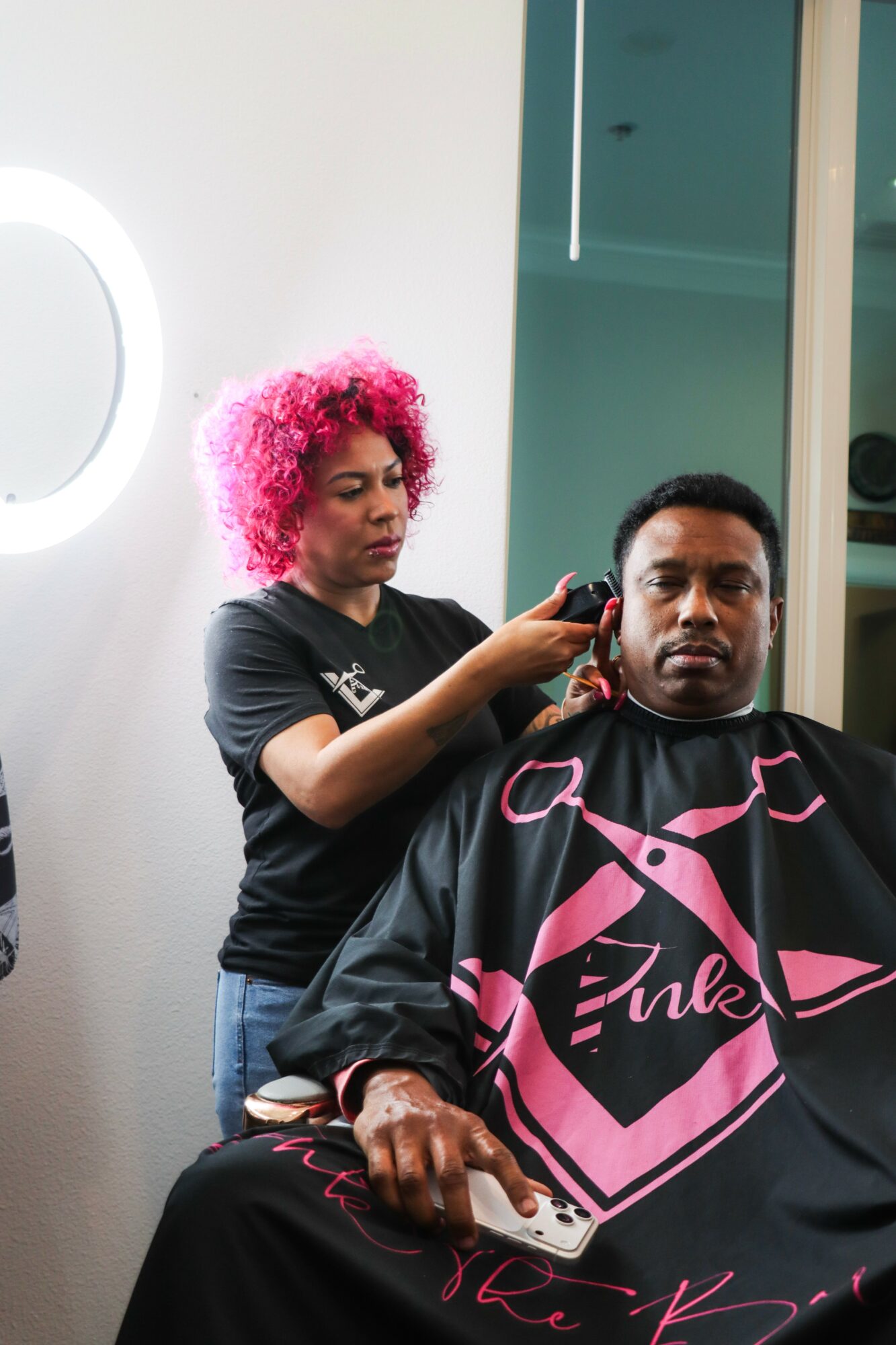 Woman with pink curly hair cutting a man's hair in a salon, man seated with a cape, salon interior visible.