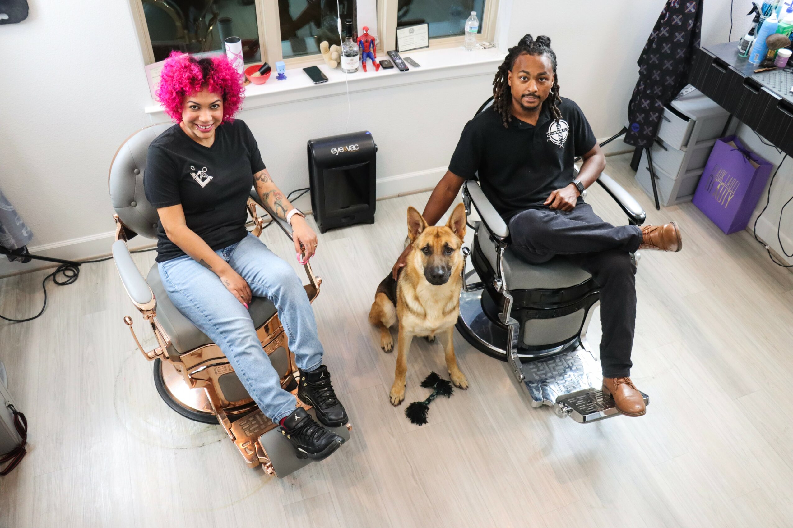 Two people and a dog in a room with light-colored flooring and a window, one person with pink hair, the other with dreadlocks.