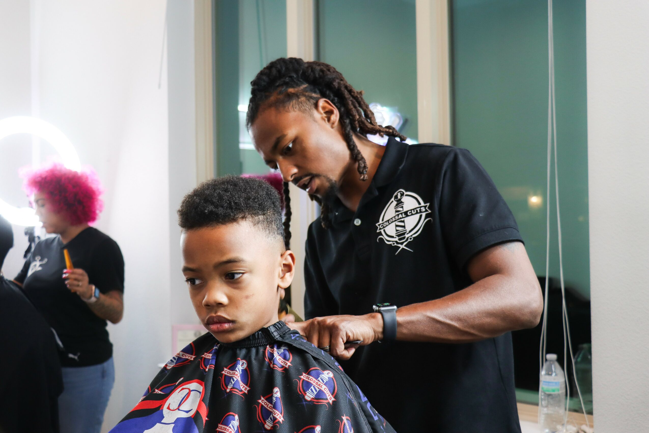 A woman with braided hair trims a young boy's hair in a salon, with another person in the background. Word logo on shirt.