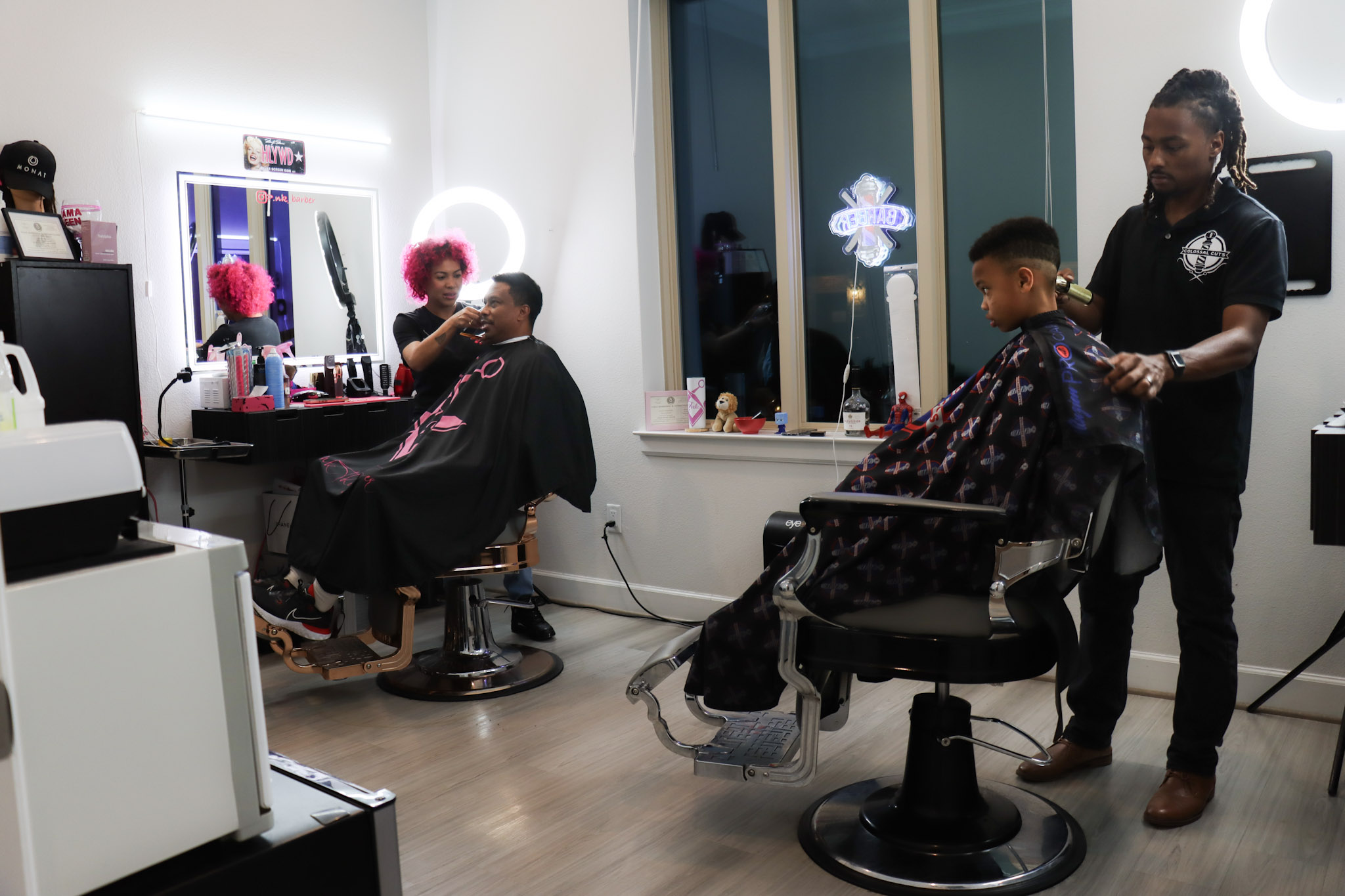 Barber cutting a child's hair while another person sits in a chair, with a stylist assisting nearby, in a salon with large windows and flowers.
