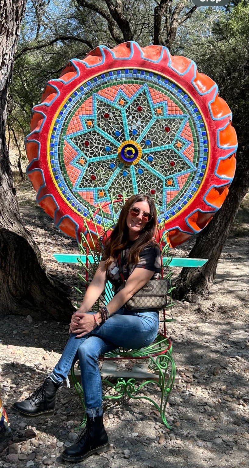 Person sitting on a green bench outdoors with a colorful decorative clock behind them, trees in the background.