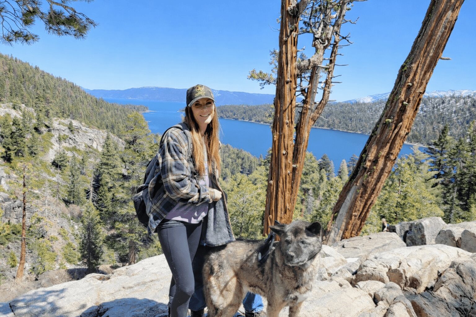 Woman with a backpack and sunglasses standing on rocks near a lake and trees, with mountains in the background.