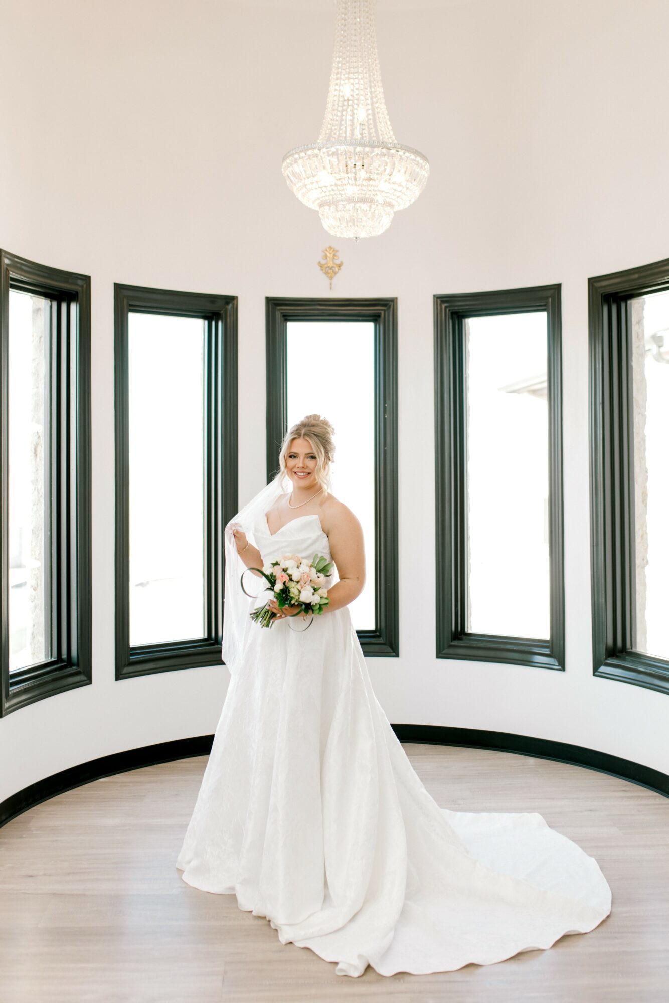 Bride in a white wedding dress holding a bouquet, standing in front of large windows with a chandelier above.