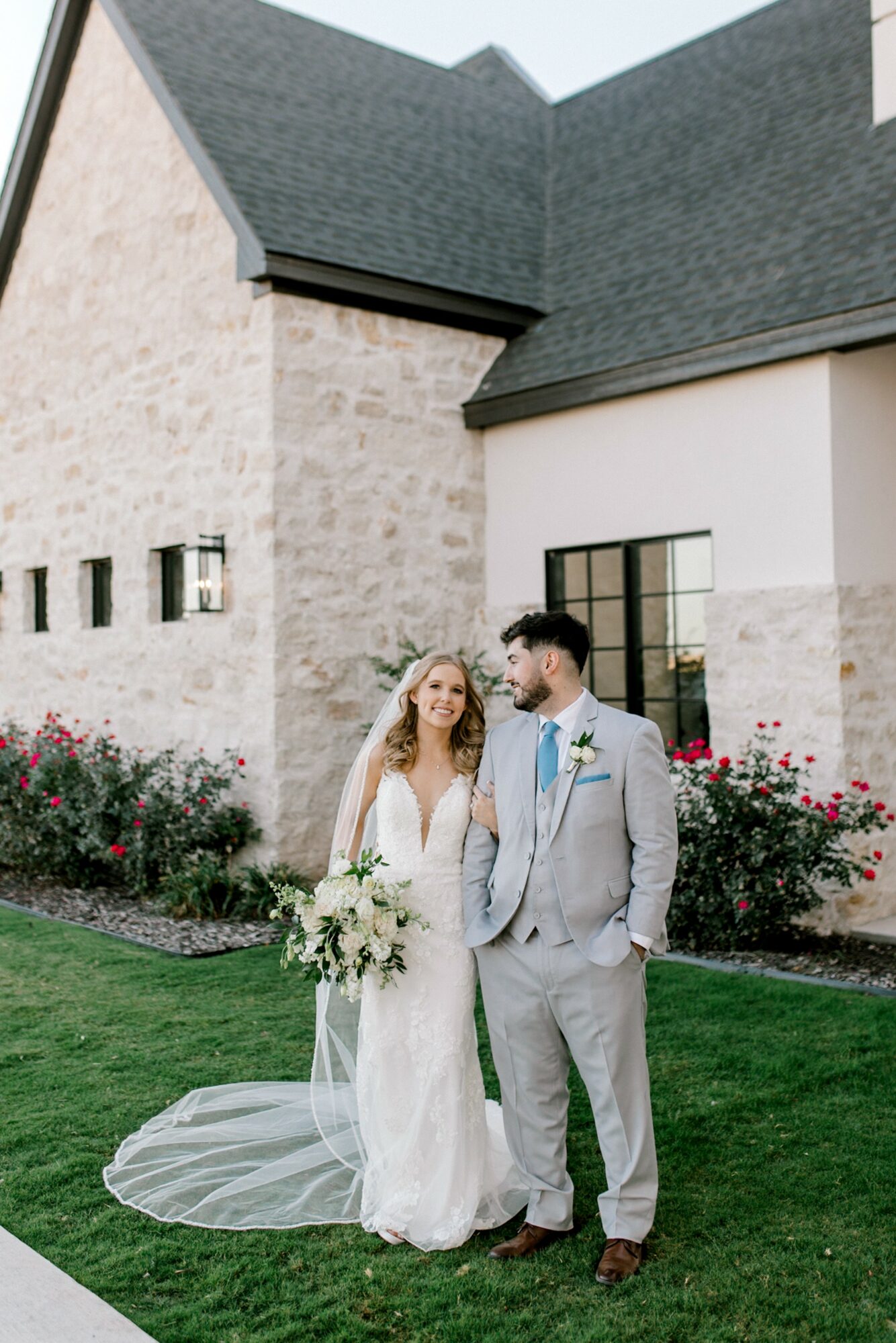Bride and groom standing outside a building with flowers, bride holding a bouquet, groom in a suit, sunny day.