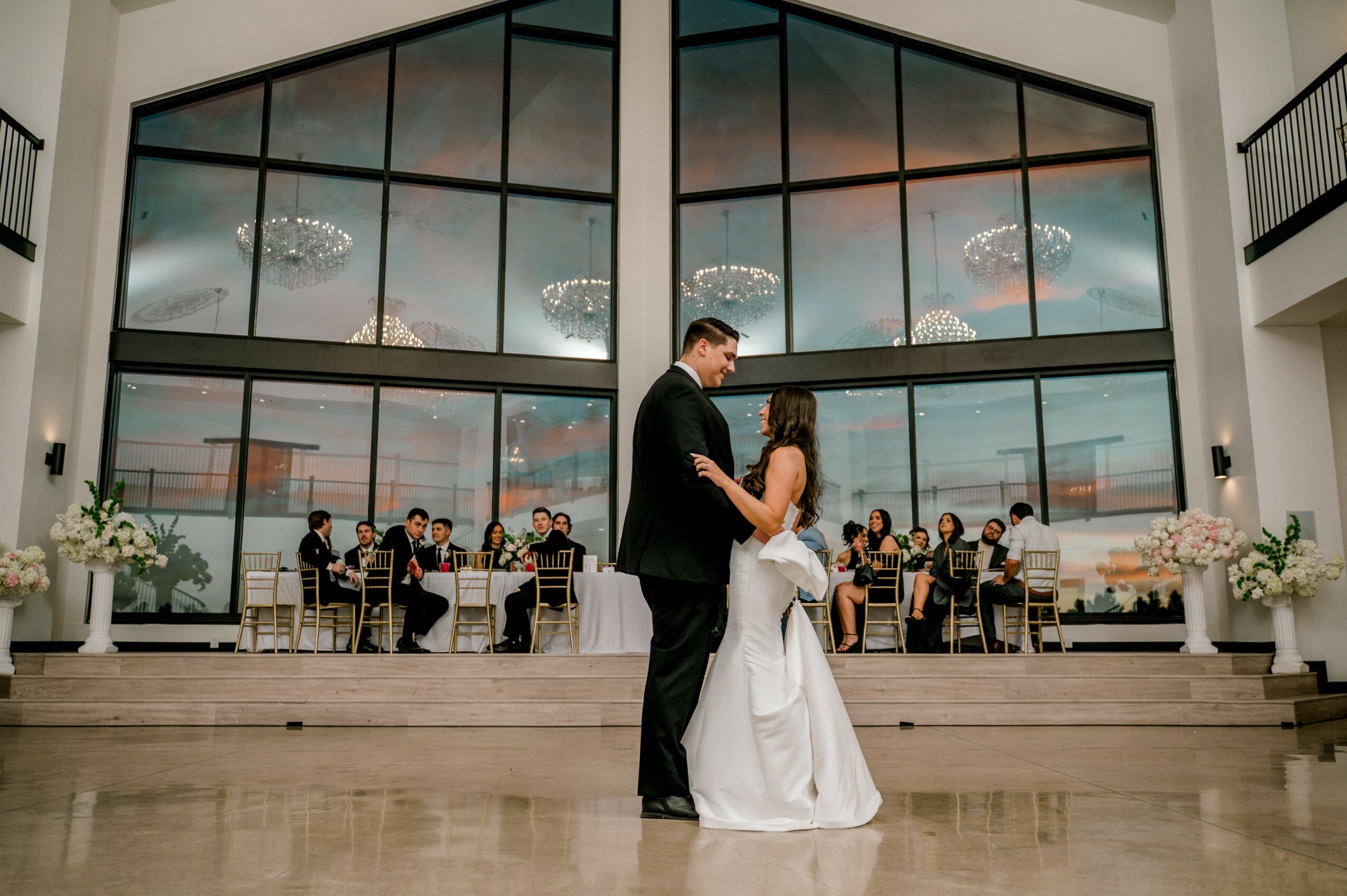 Couple dancing in a spacious hall with large windows and a city view, guests seated at tables in the background.