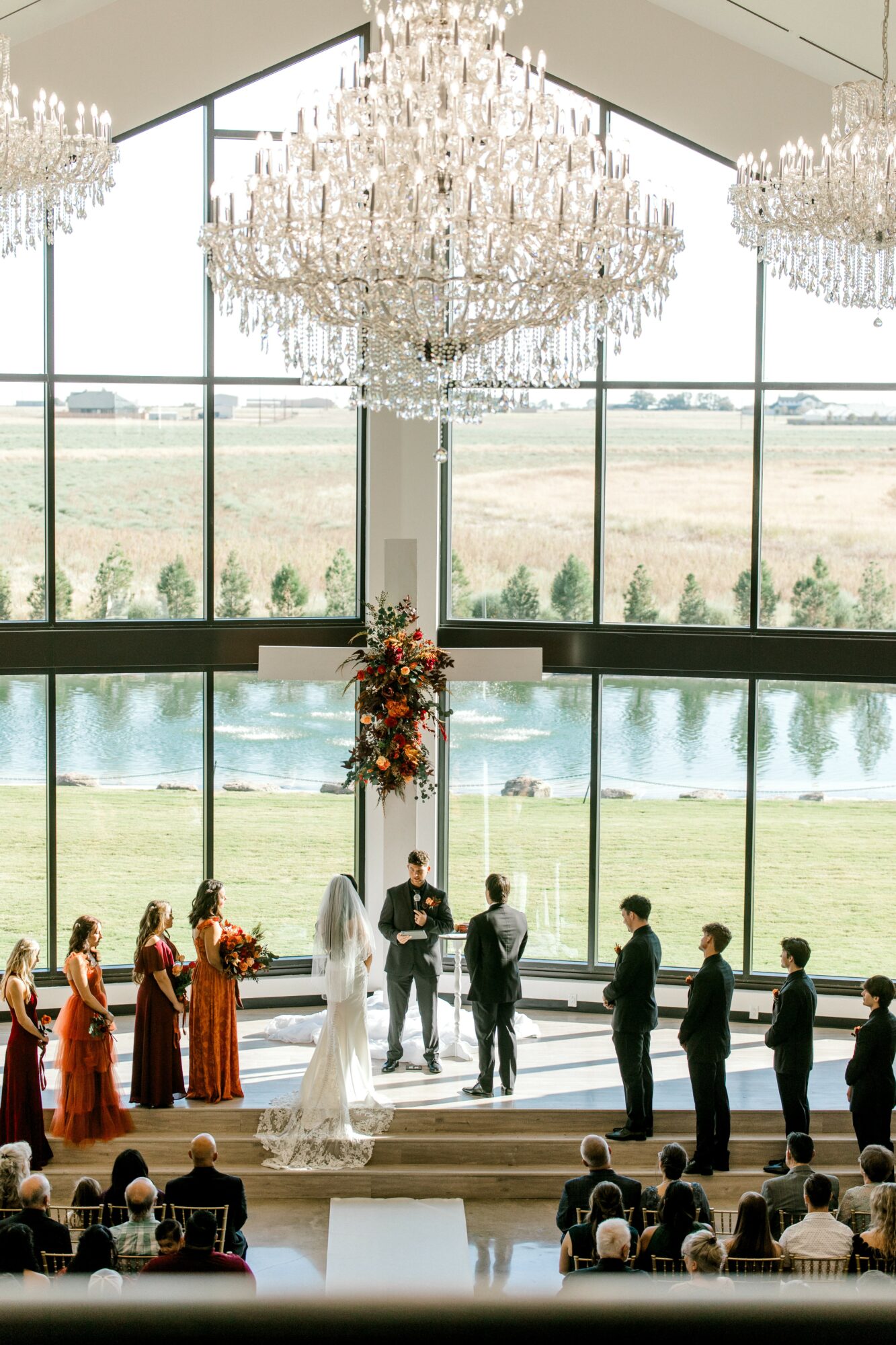 Wedding ceremony with a bride, groom, bridesmaids, groomsmen, and guests inside a bright venue with large windows and chandeliers.