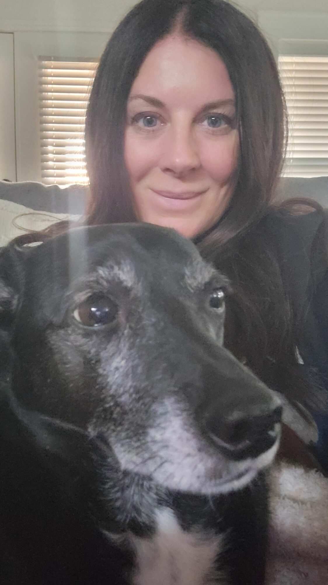 Woman with dark hair smiling next to a large black and white dog indoors, window with blinds in background.