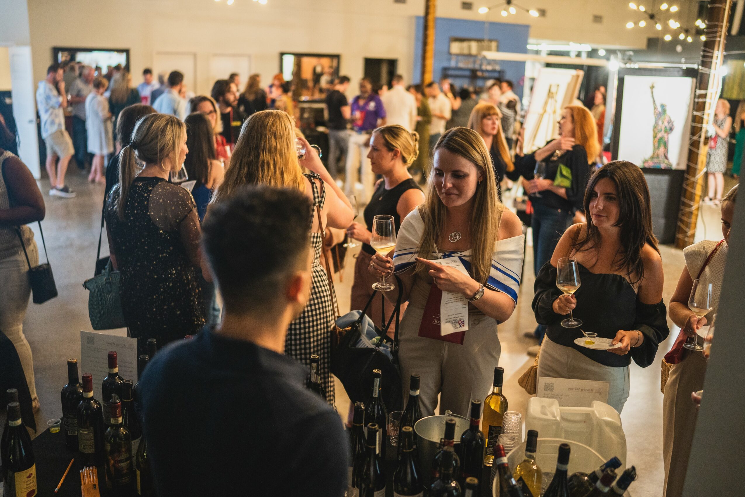People gather at a busy indoor event, some holding drinks, with booths and displays in the background.