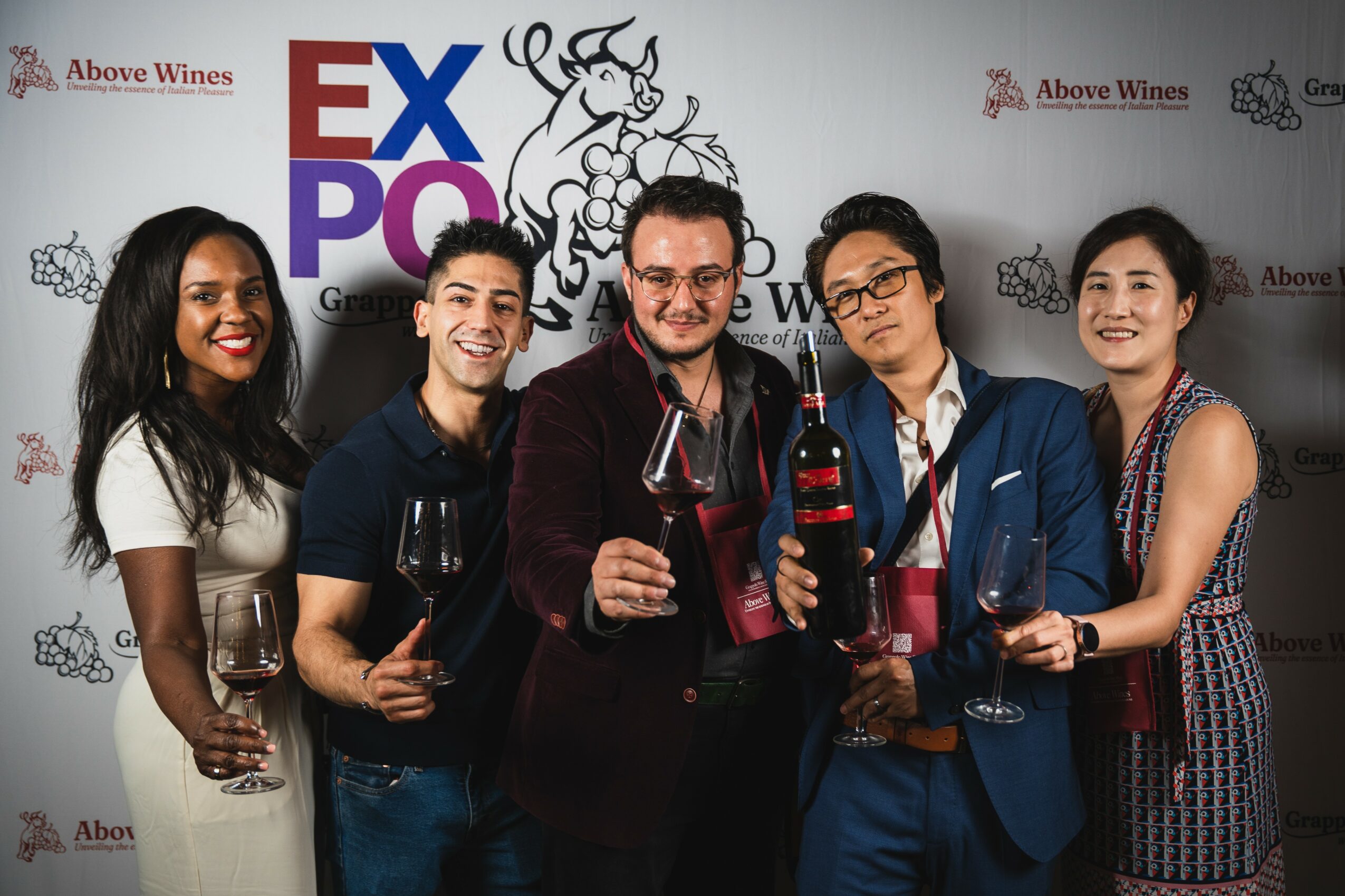 Five people smiling and holding wine glasses at an event, standing in front of a backdrop with logos and text.