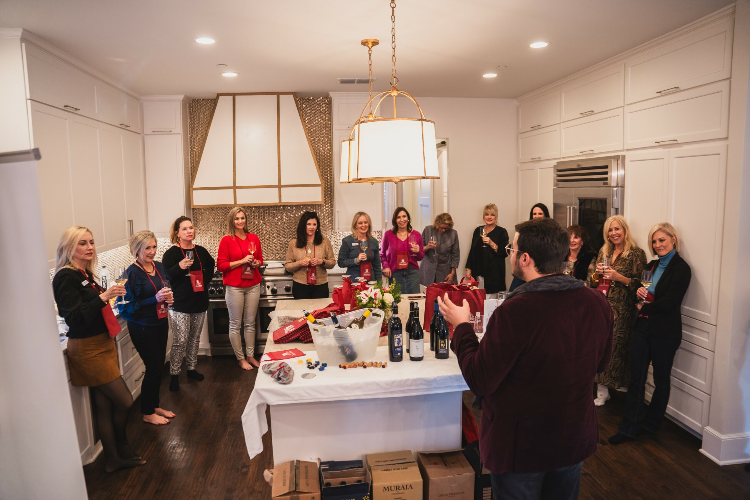 Group of women gathered in a bright, elegant kitchen around a table with drinks and gifts, celebrating together.