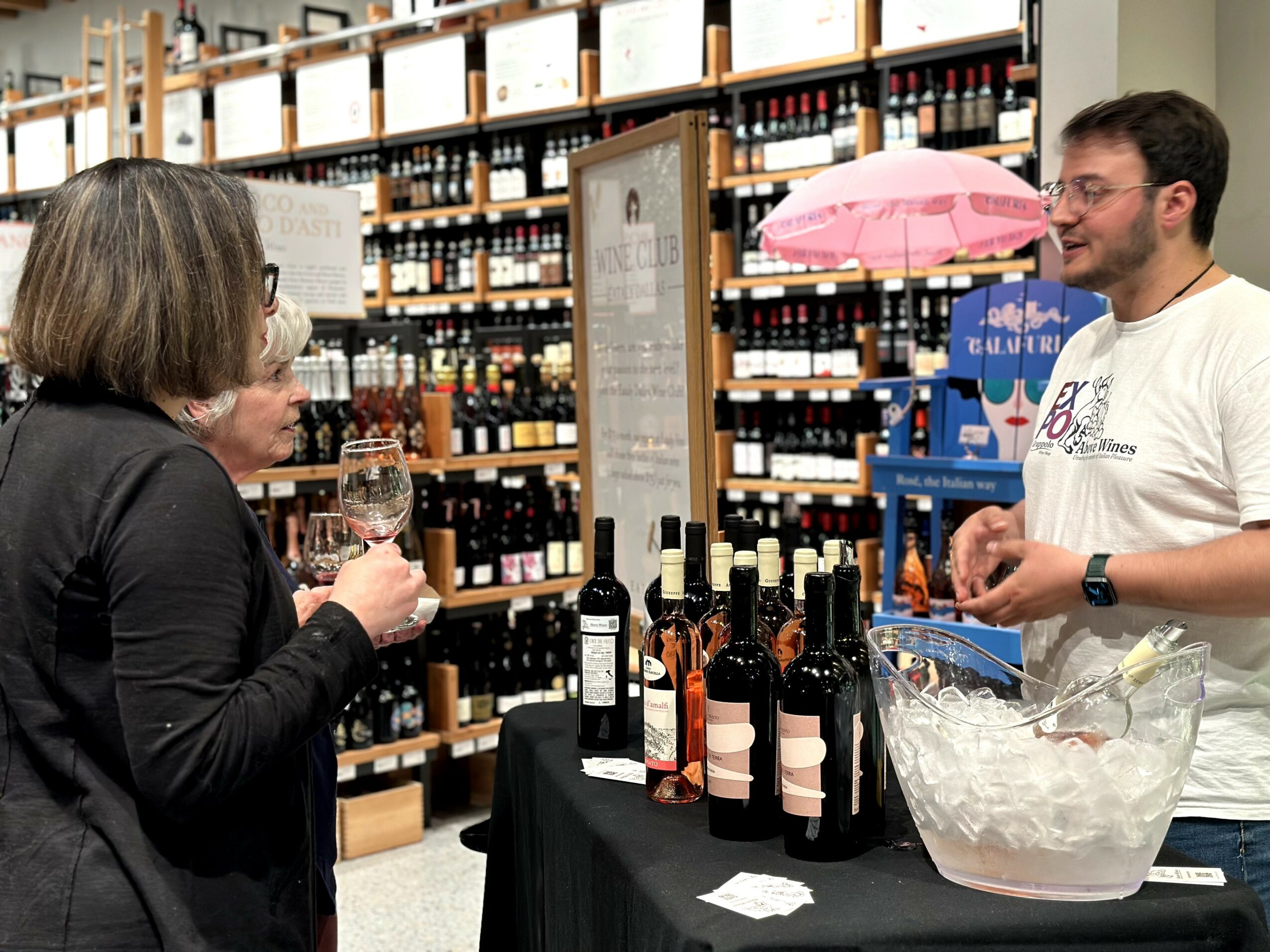 A woman with glasses and a man with a beard converse at a wine tasting booth in a store, with shelves of bottles behind them.