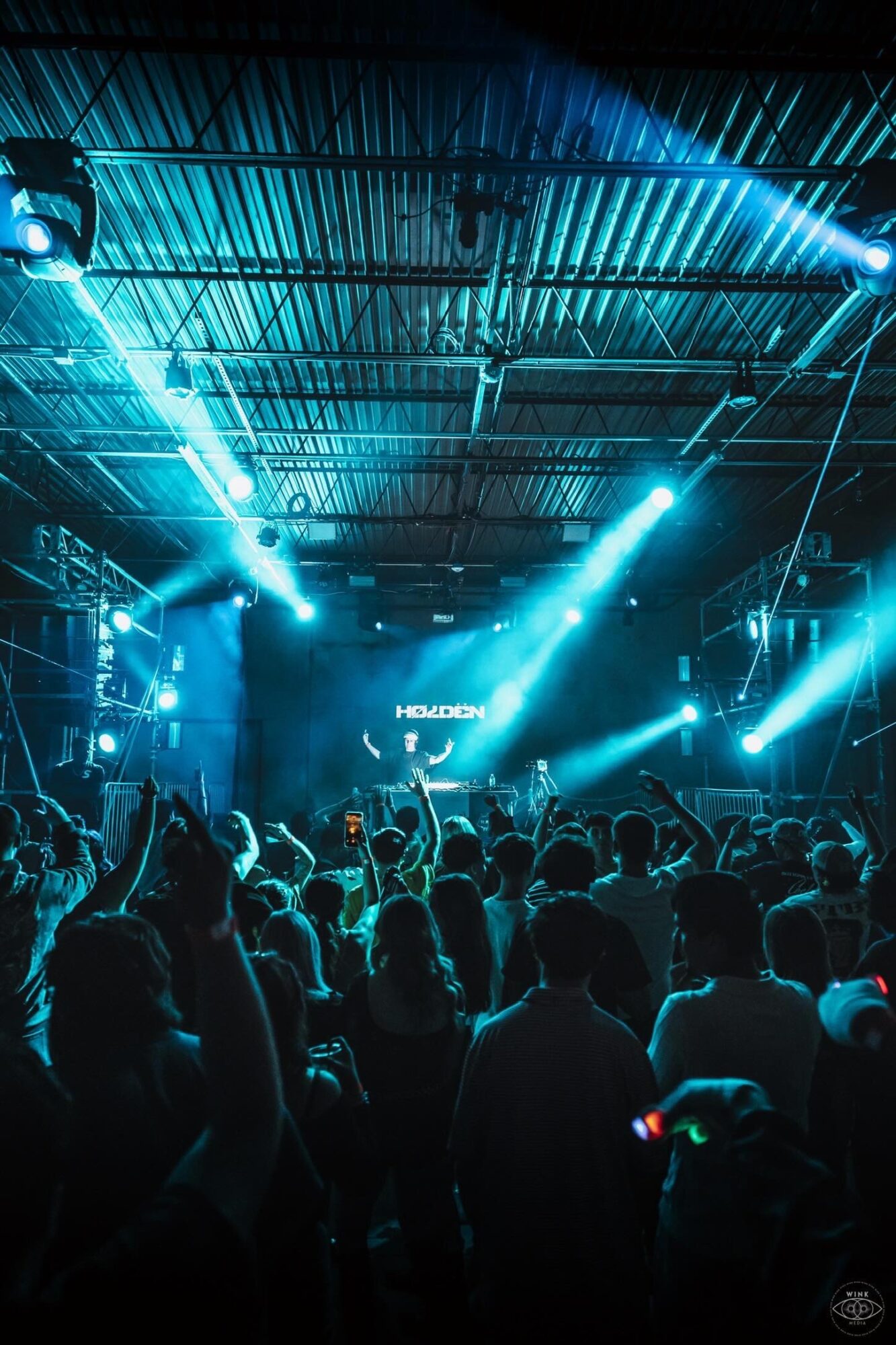 Crowd at concert with bright blue stage lights and a DJ booth, audience facing stage, dark indoor venue.