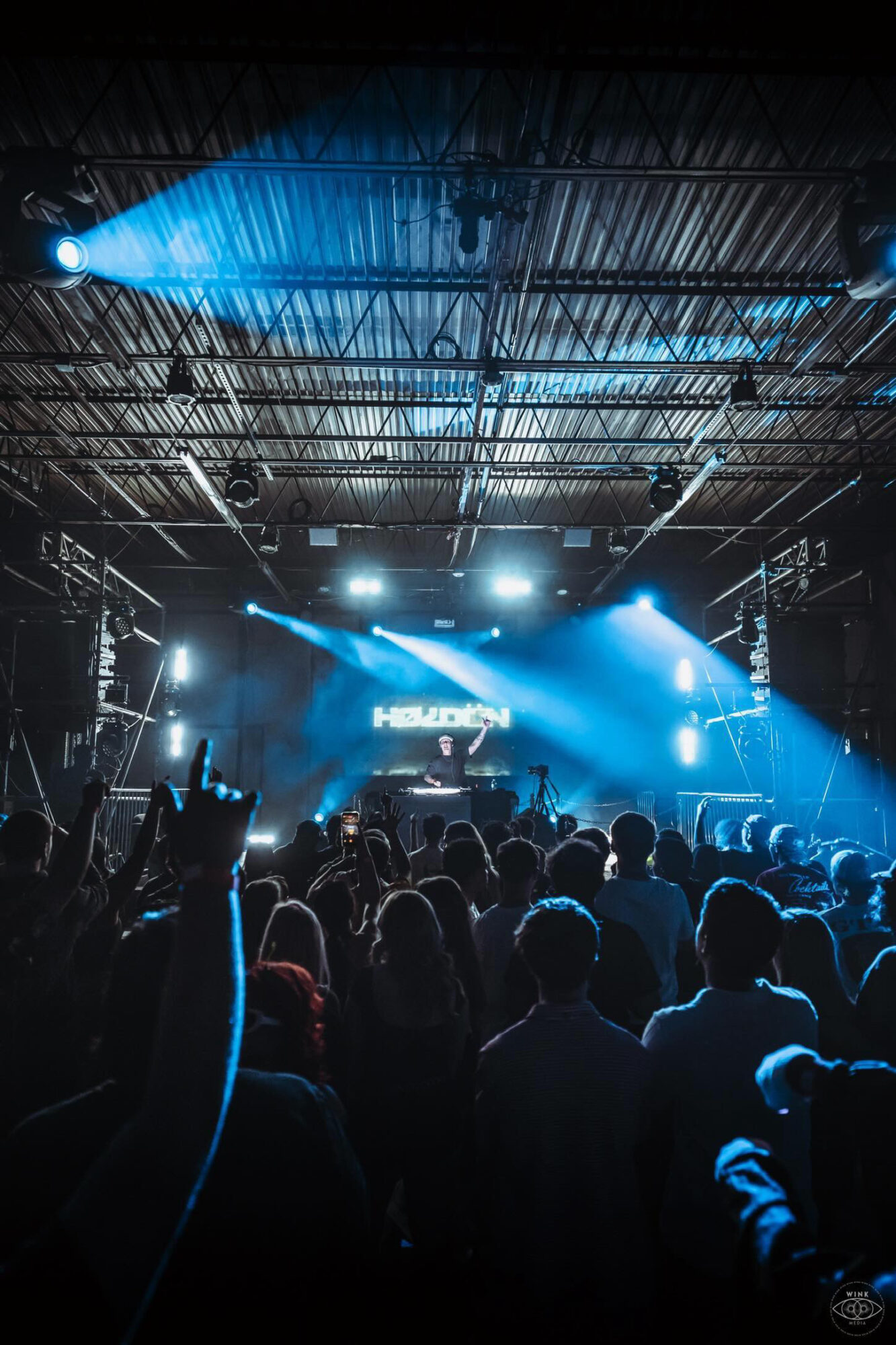 Crowd at concert with stage lights and performers, audience raising hands, dark indoor venue, vibrant blue lighting.