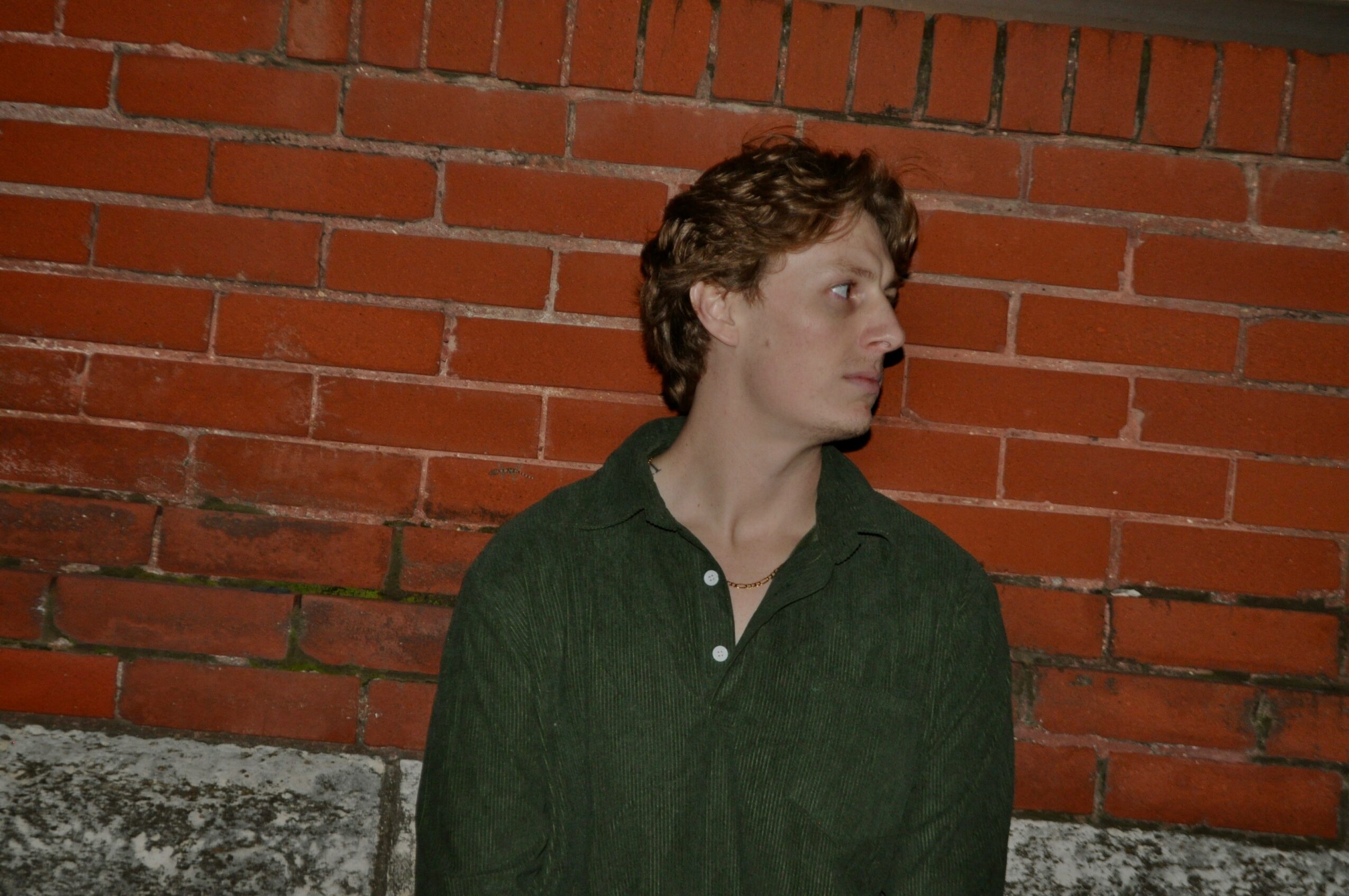 Young man with curly hair wearing a dark green shirt, standing against a brick wall, looking to the side.