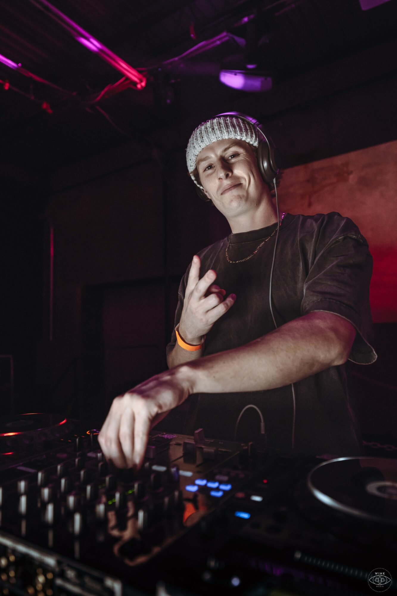 DJ wearing headphones and a striped beanie, making a hand gesture in a dark, colorful club setting.