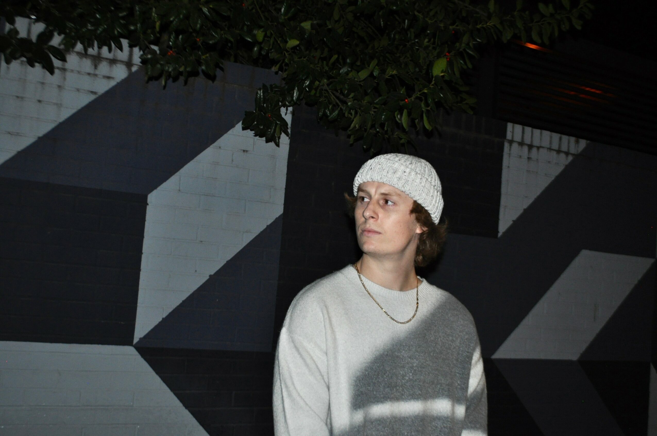 Person wearing a white knit hat and sweater stands outdoors near a geometric black and white wall, looking to the side.