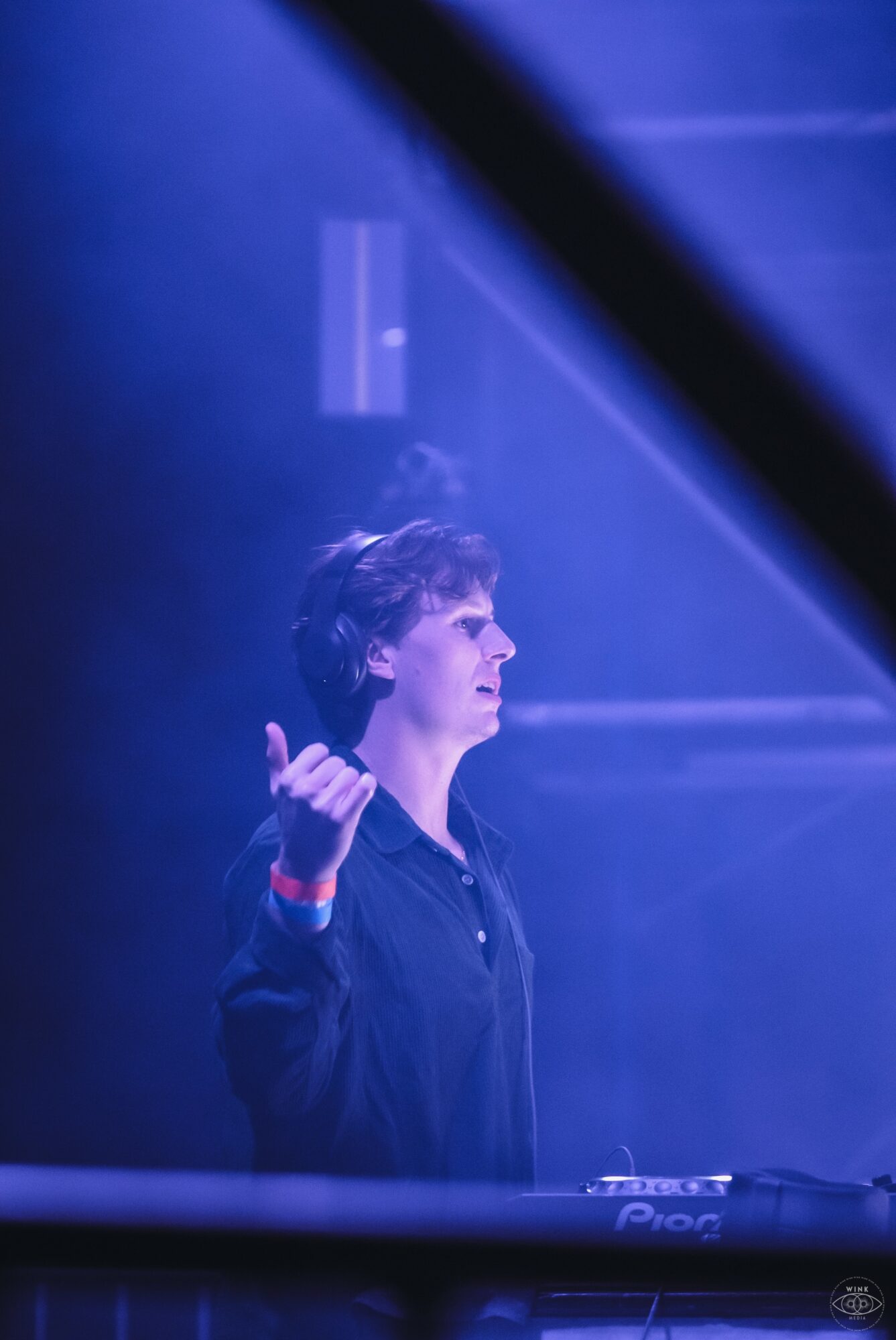 Person with short hair standing in blue-lit environment, facing right, with hand raised, possibly on stage.