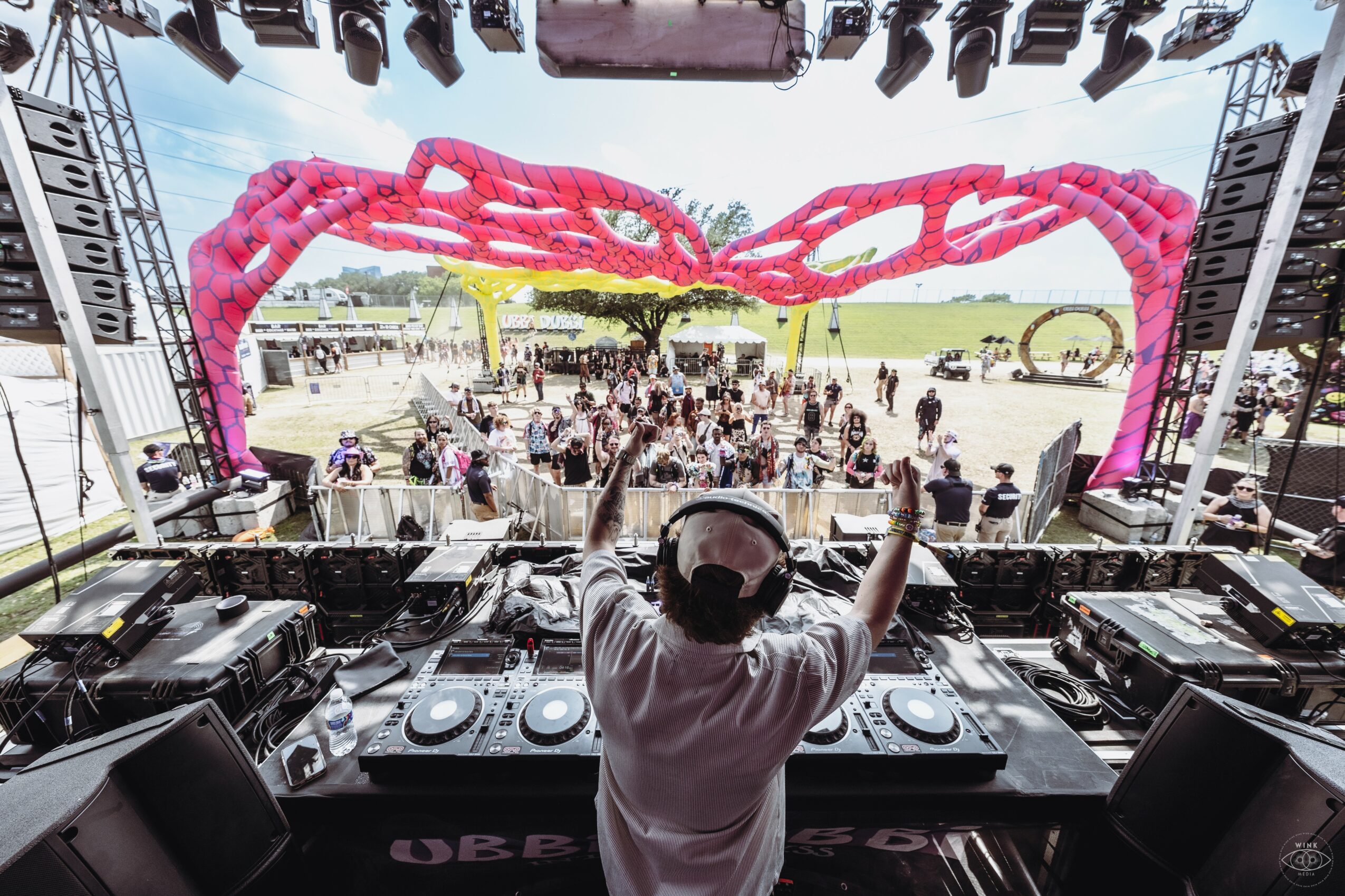 DJ performing at an outdoor event with a large pink balloon sculpture overhead, crowd in the background, stage setup visible.