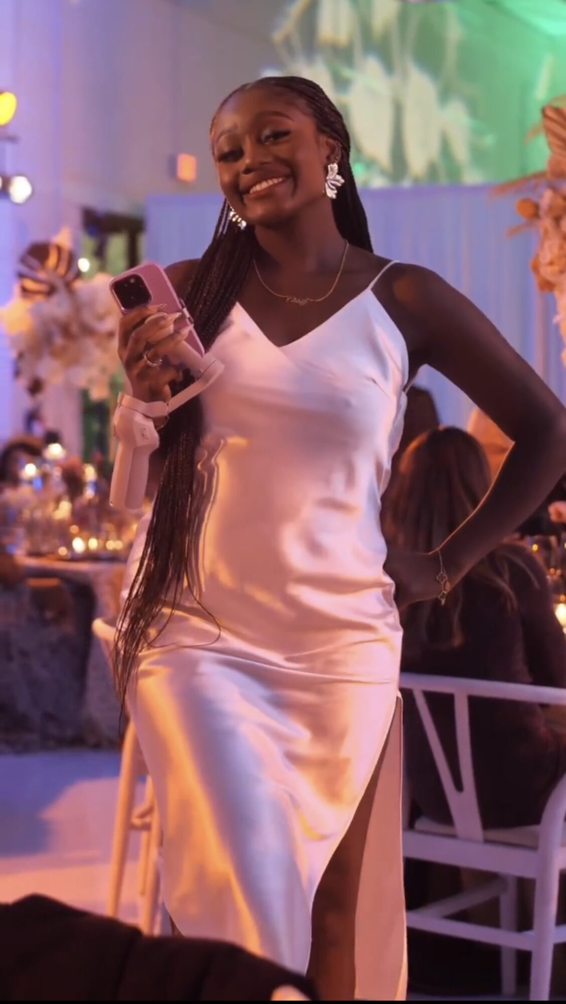 Smiling woman in white dress holding a phone at an event with colorful decorations.