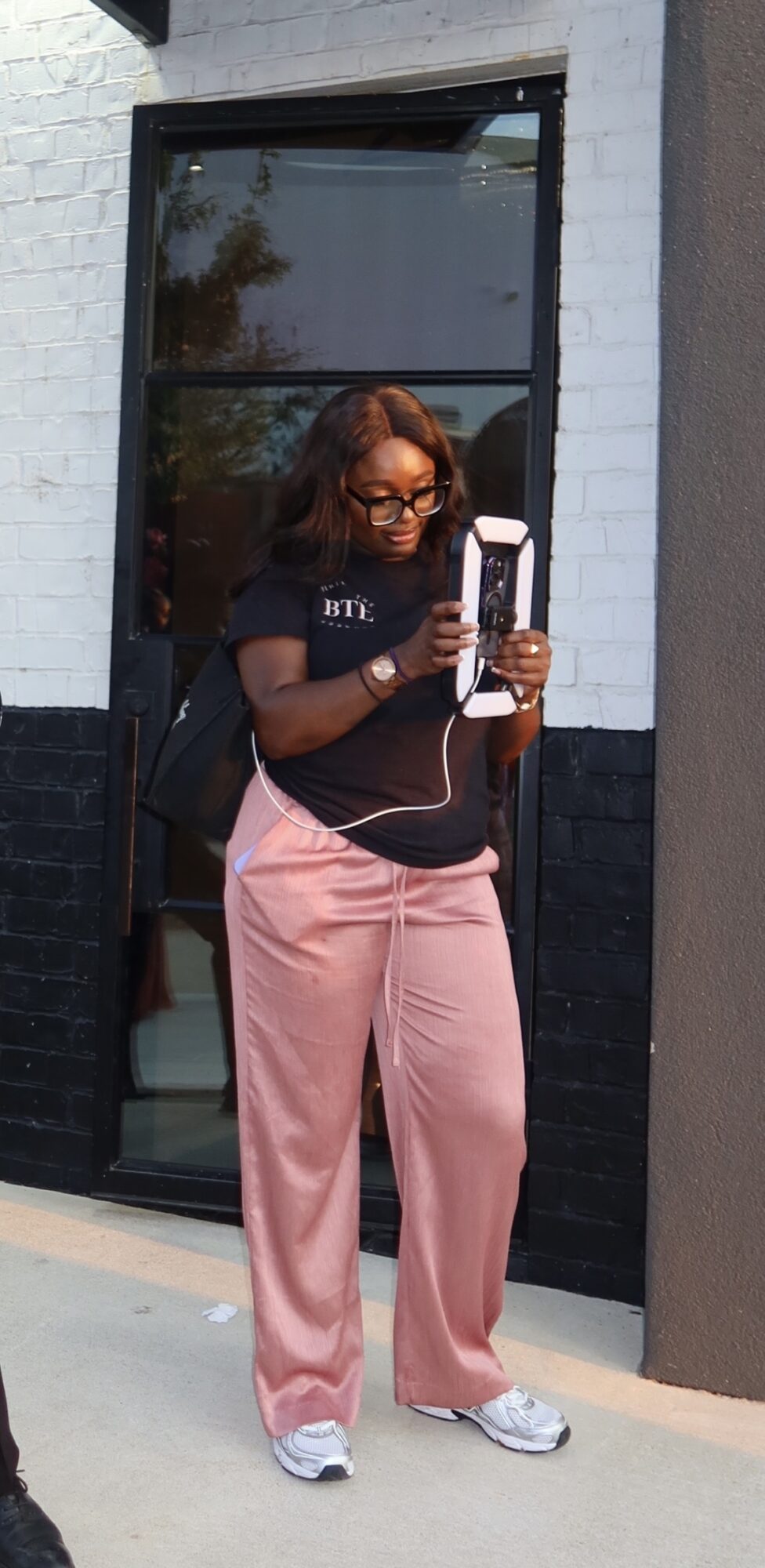 Woman with curly hair wearing glasses, black T-shirt, pink pants, and white sneakers, holding a phone outside a building.