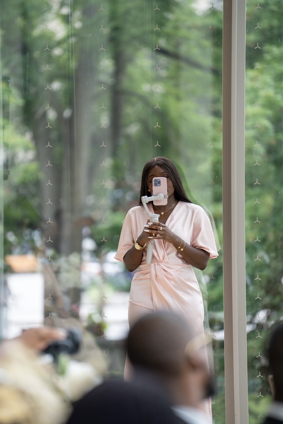 Person taking a mirror selfie with a smartphone, wearing a light-colored dress, in front of a large window with green trees outside.