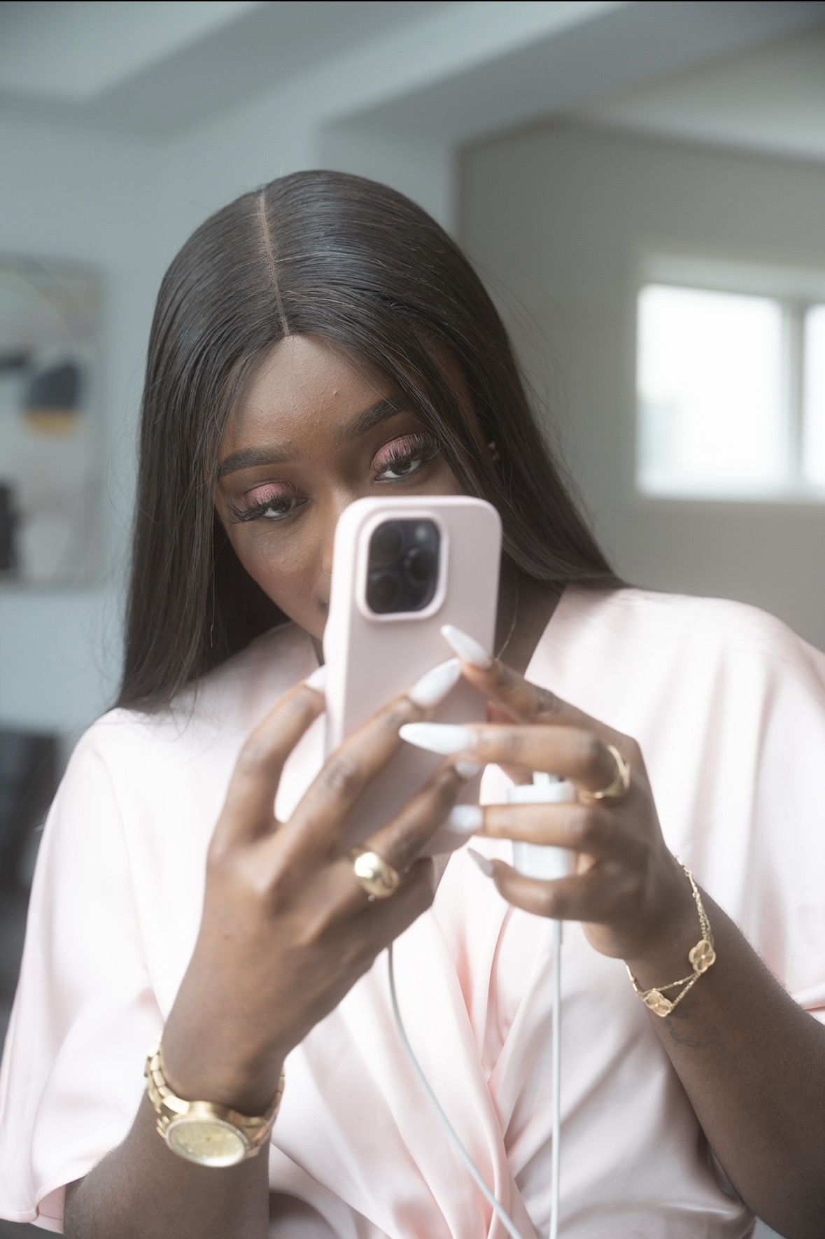 Woman with long dark hair taking a selfie with a white smartphone, wearing a white top and gold jewelry.