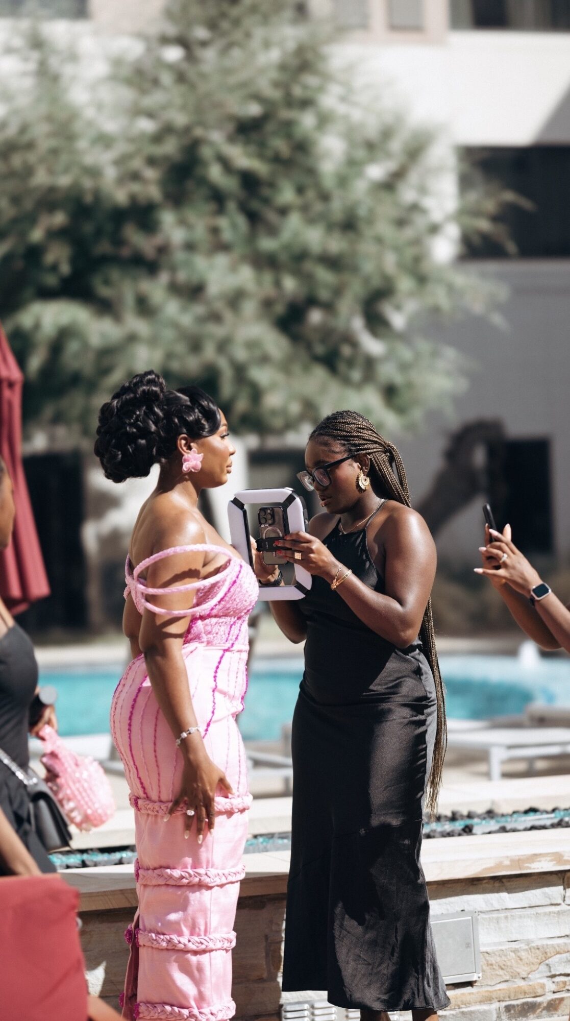 Two women standing outdoors near a pool, one reading a phone, the other wearing sunglasses and a pink dress, with trees and a building in background.