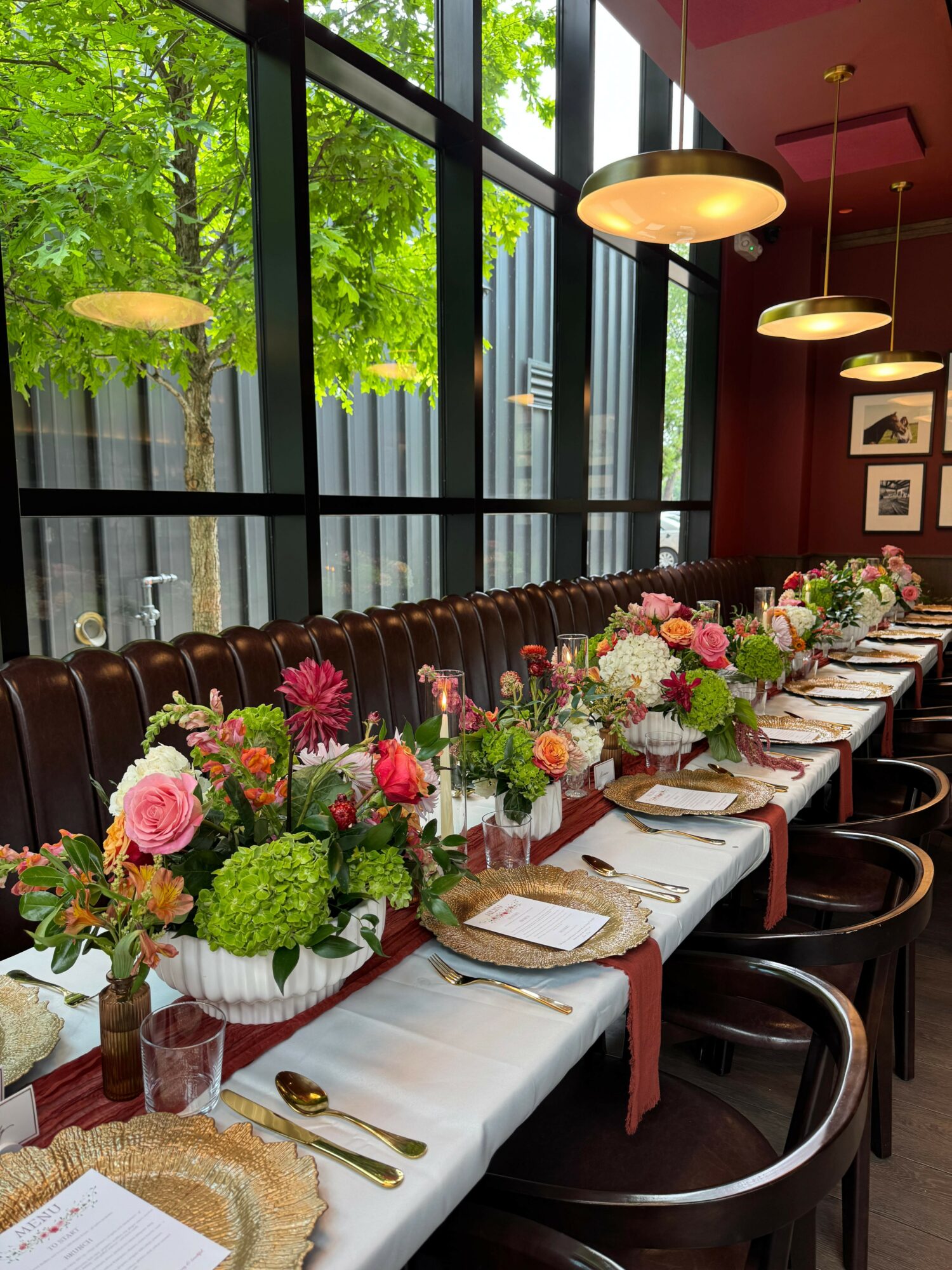 Long dining table with floral arrangements, white tablecloths, and place settings inside a room with large windows and hanging lights.