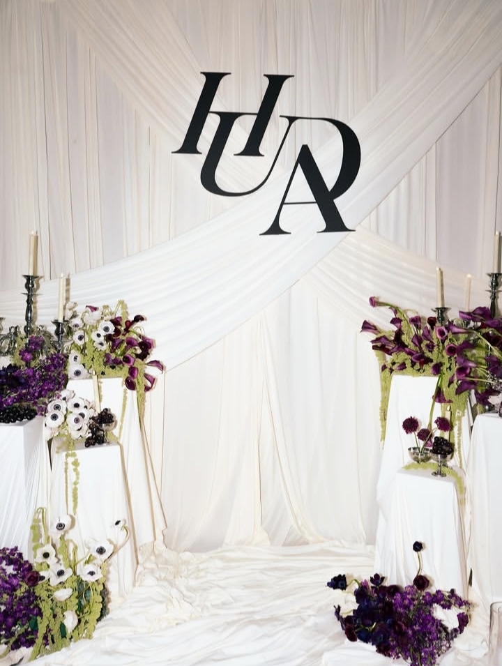 Decorative backdrop with large black letters 'H', 'U', and 'A' above white draped fabric, floral arrangements on sides.