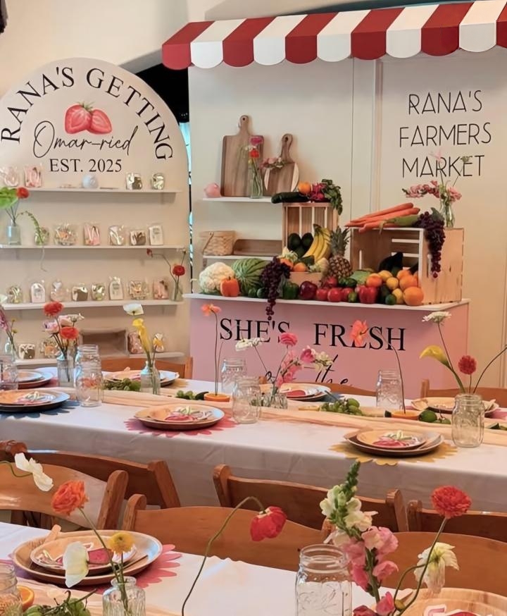Indoor farmers market display with shelves of jars, fresh flowers, and a pink sign reading 'She's Fresh'.