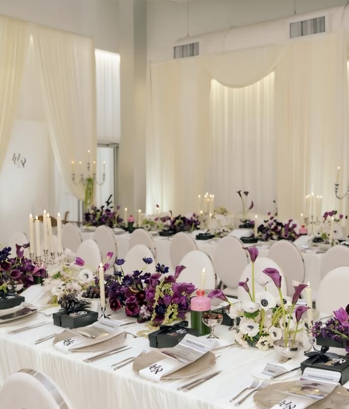 Elegant banquet table with white tablecloths, floral centerpieces, and place settings in a bright, airy room with white curtains.