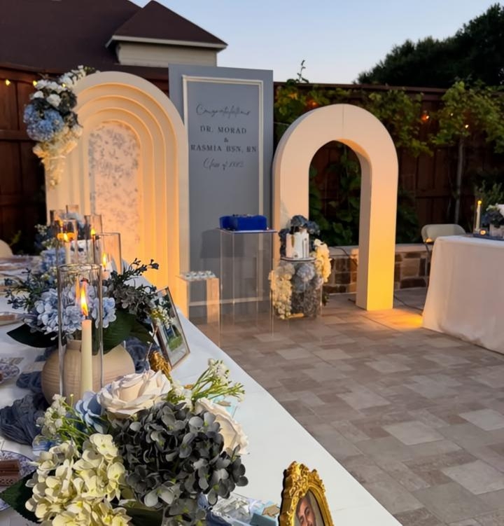 Outdoor celebration setup with decorative arches, floral arrangements, and a sign, on a paved patio with a house and trees in background.
