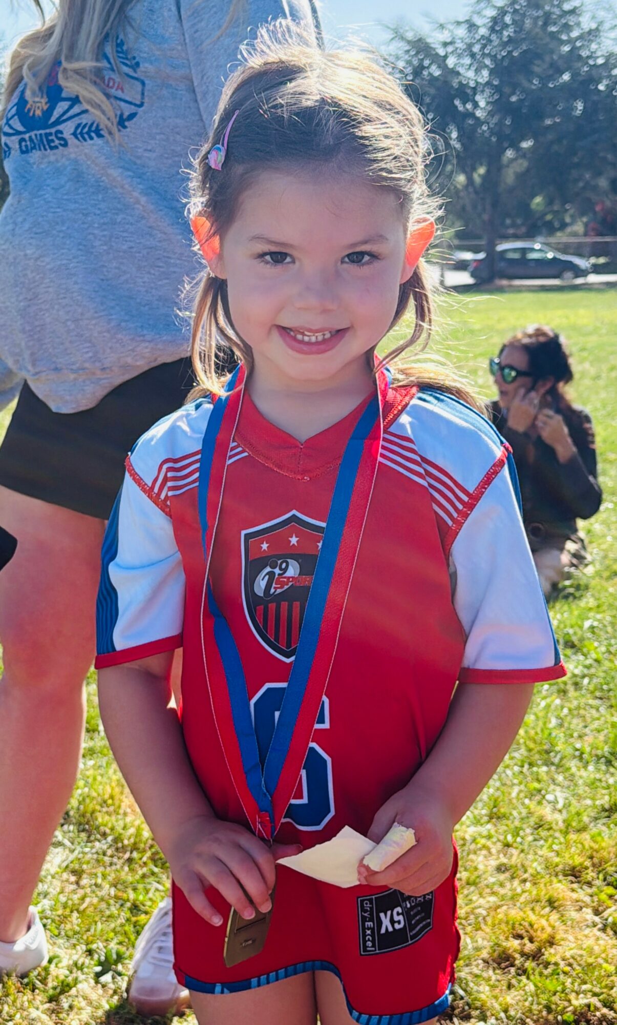 Young girl with a medal around her neck, smiling outdoors in a red and white sports jersey, with other children and trees in background.